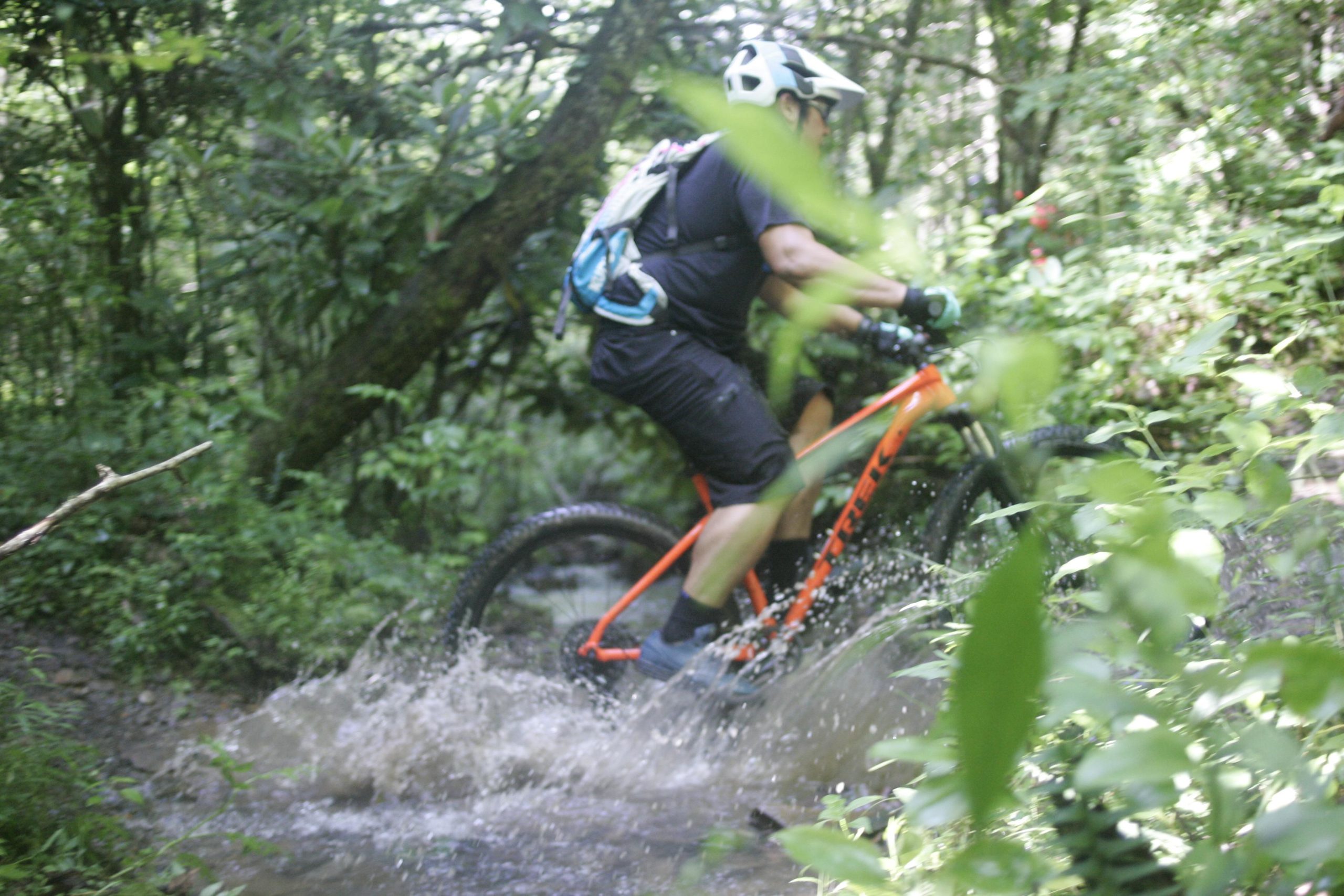 A person riding a mountain bike through a forested area, splashing through a muddy puddle. The cyclist is wearing a helmet and a backpack, surrounded by lush greenery and trees. Tsali Left Loop mountain bike trail.