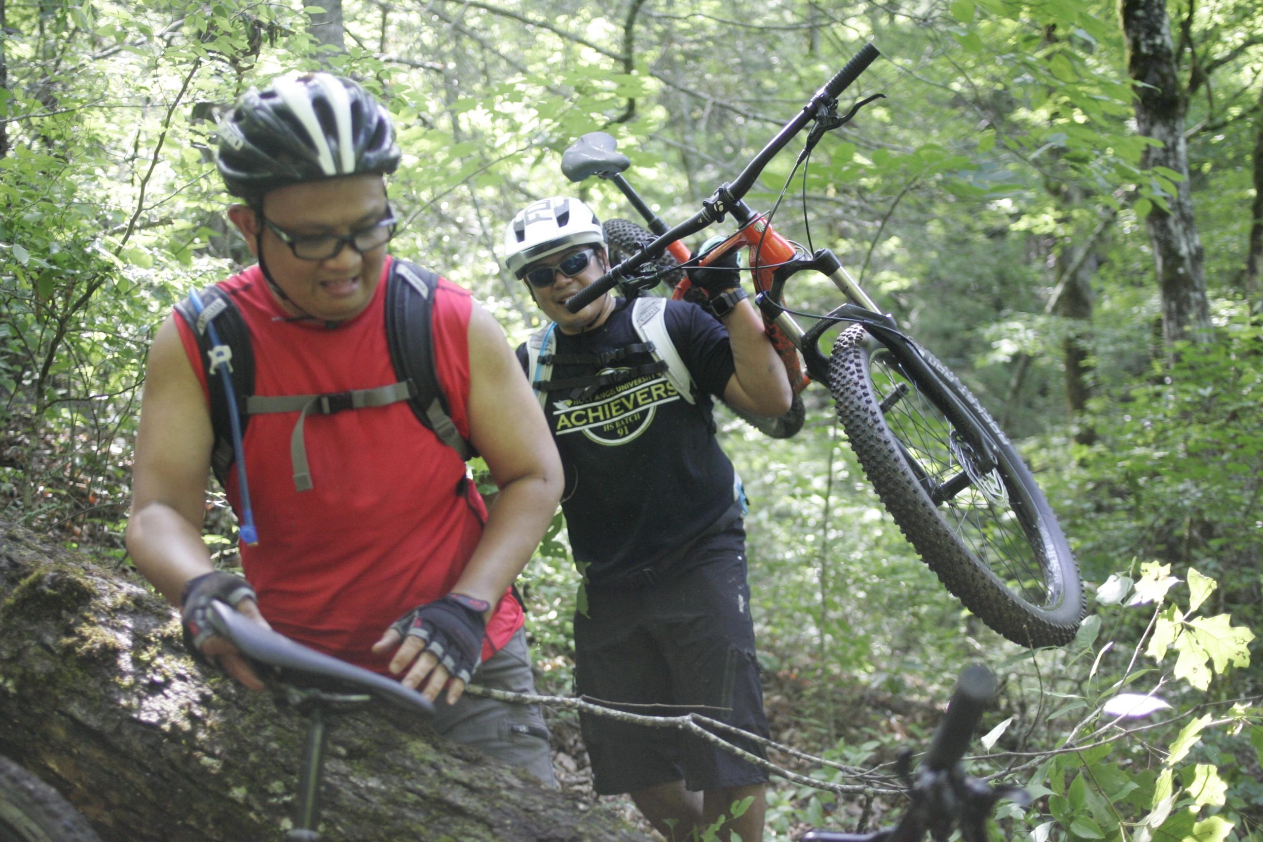 Two mountain bikers navigating a forest trail. One is wearing a red sleeveless shirt and glasses, looking at the ground, while the other, in a black shirt and helmet, is smiling and carrying a mountain bike over his shoulder. The scene is lush with green foliage, indicating a summer day. Tsali Left Loop mountain bike trail.
