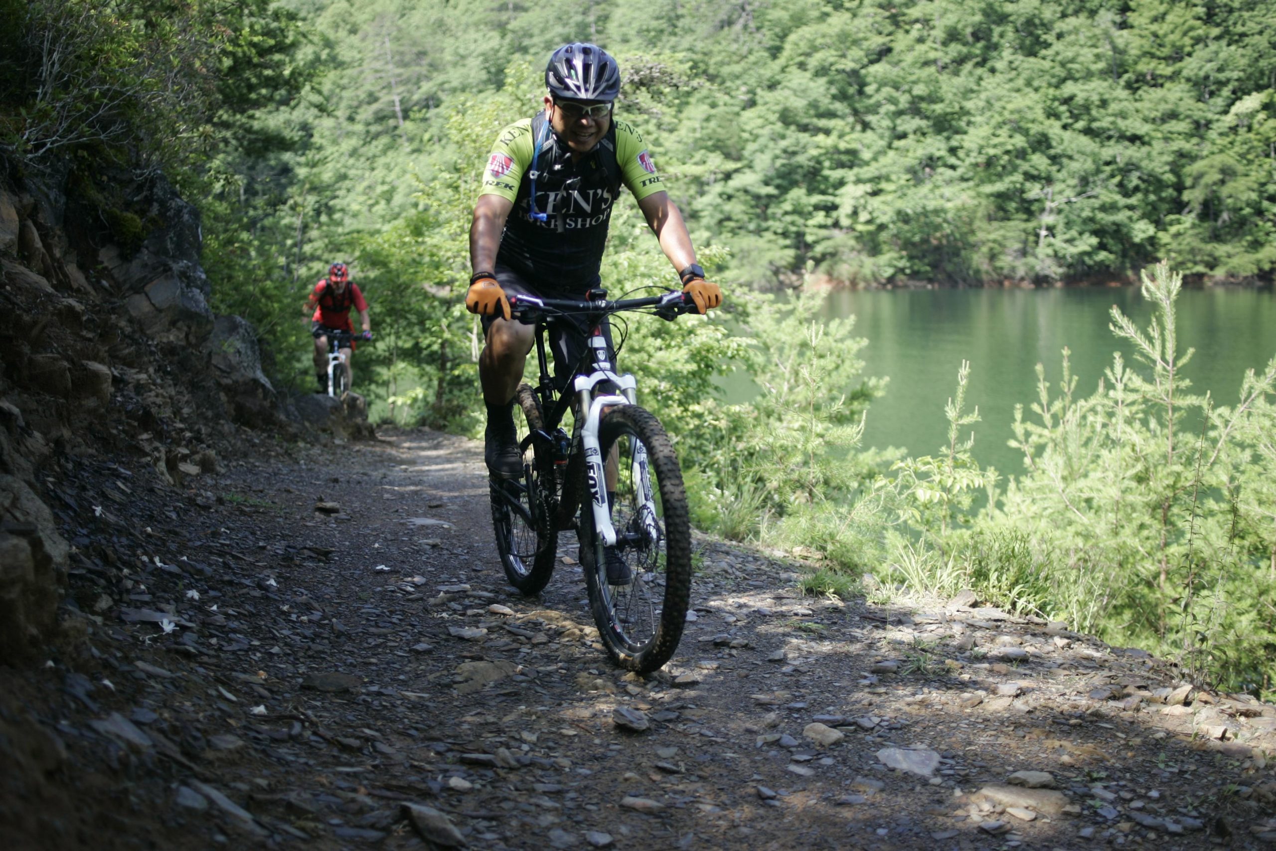 Two mountain bikers riding on a rocky trail near a lake surrounded by greenery. One biker, wearing a black and green jersey and yellow gloves, is in the foreground, while another biker in red is further back. The scene is sunny, showcasing the vibrant nature around them. Tsali Left Loop mountain bike trail.