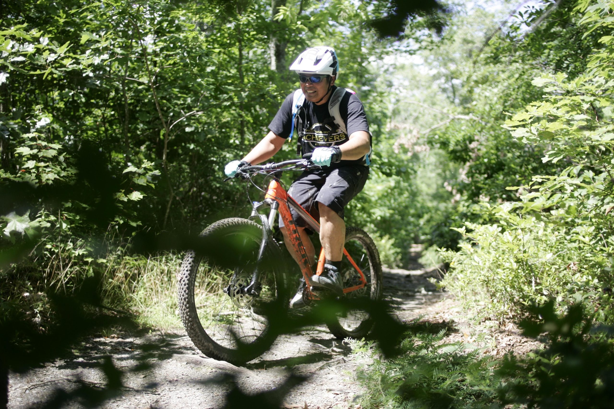 A person riding a mountain bike on a dirt trail surrounded by green foliage. The rider is wearing a helmet, sunglasses, and gloves, and is smiling as they navigate through the natural landscape. Tsali Left Loop mountain bike trail.