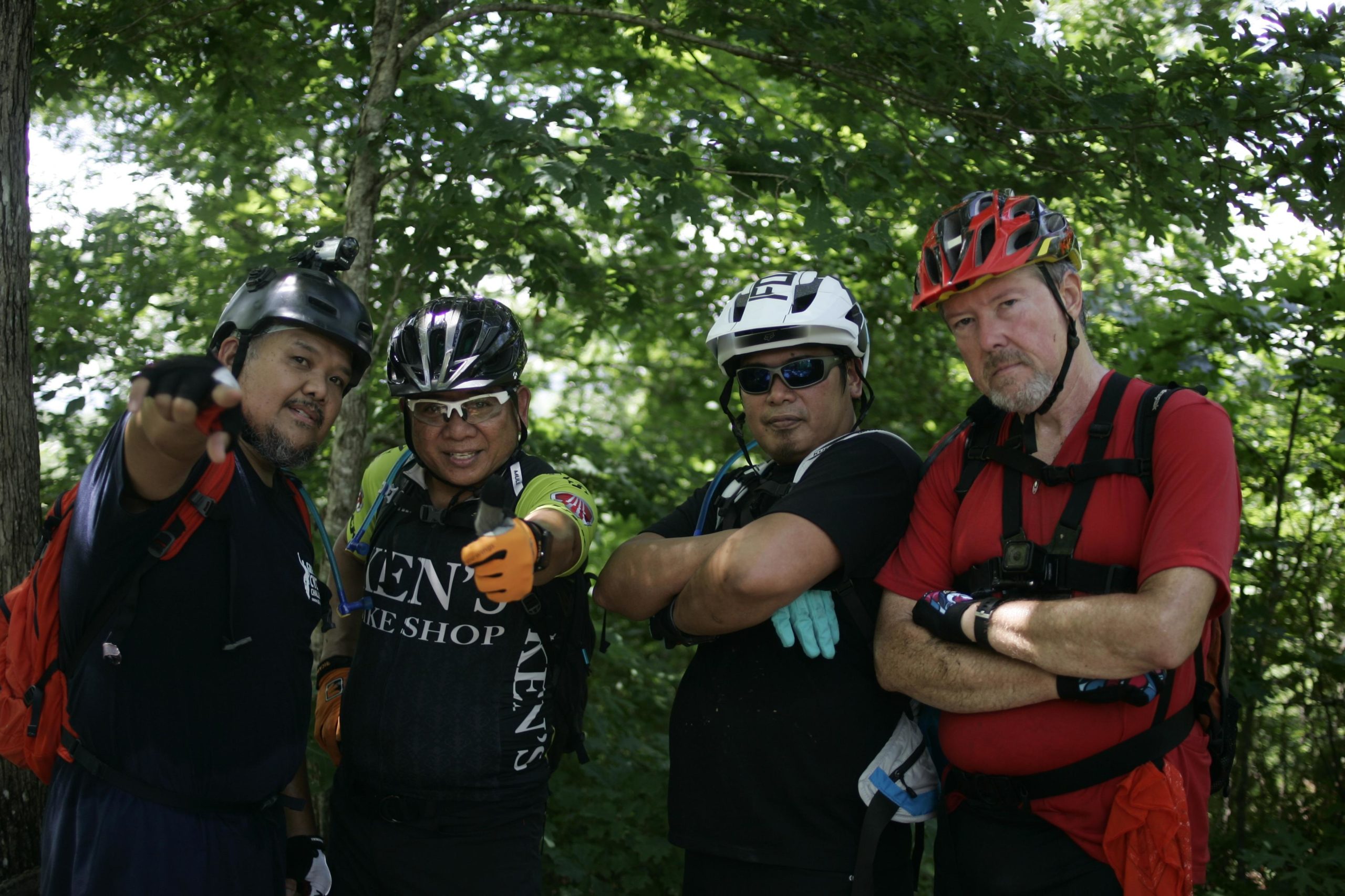 Four mountain bikers pose for a photo in a forested area, wearing helmets and protective gear. Two of them are giving thumbs up, while the others have their arms crossed. They are surrounded by lush green trees, suggesting an outdoor adventure setting. Tsali Left Loop mountain bike trail.