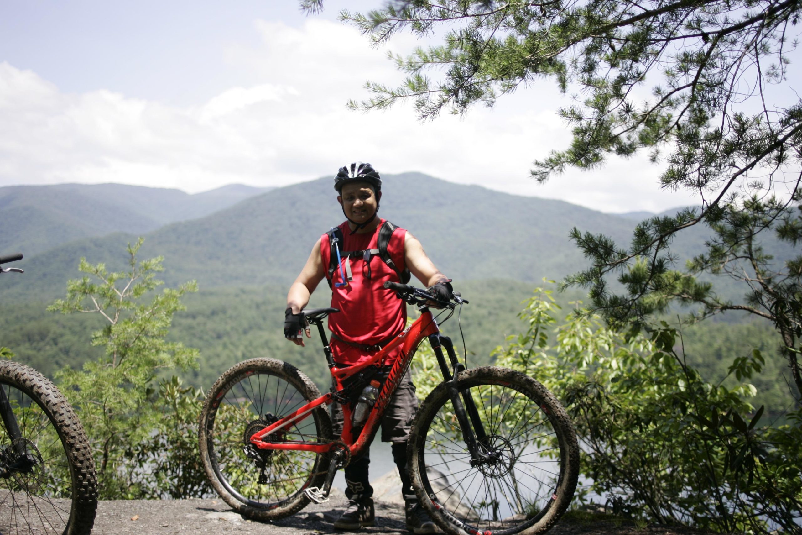 A person wearing a red sleeveless shirt and a bike helmet stands next to a red mountain bike with muddy tires on a rocky outcrop. The background features lush green mountains and a cloudy blue sky. Tsali Left Loop mountain bike trail.