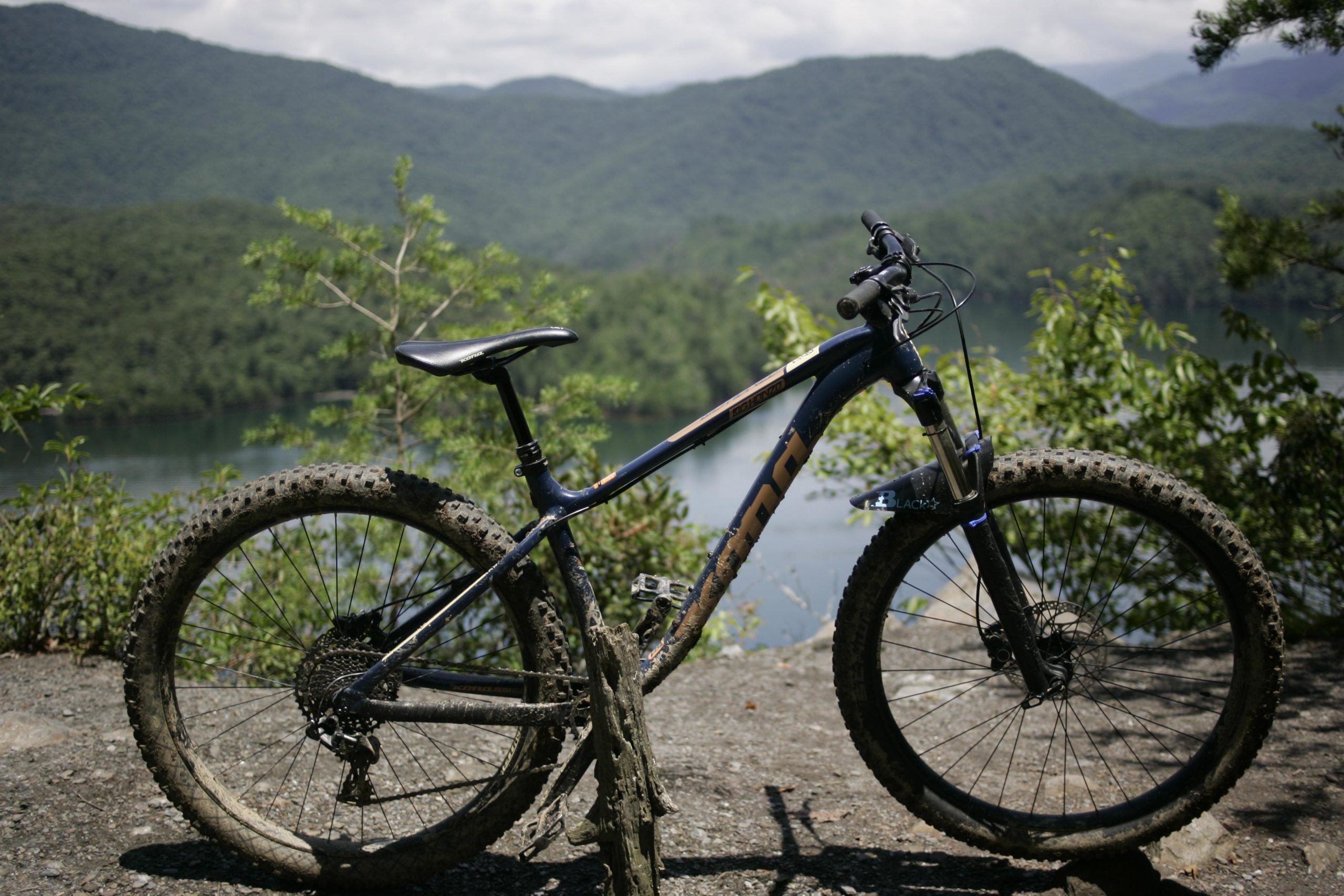 A mountain bike with muddy tires rests on a rocky surface, overlooking a serene lake surrounded by green mountains and trees under a partly cloudy sky. Tsali Left Loop mountain bike trail.