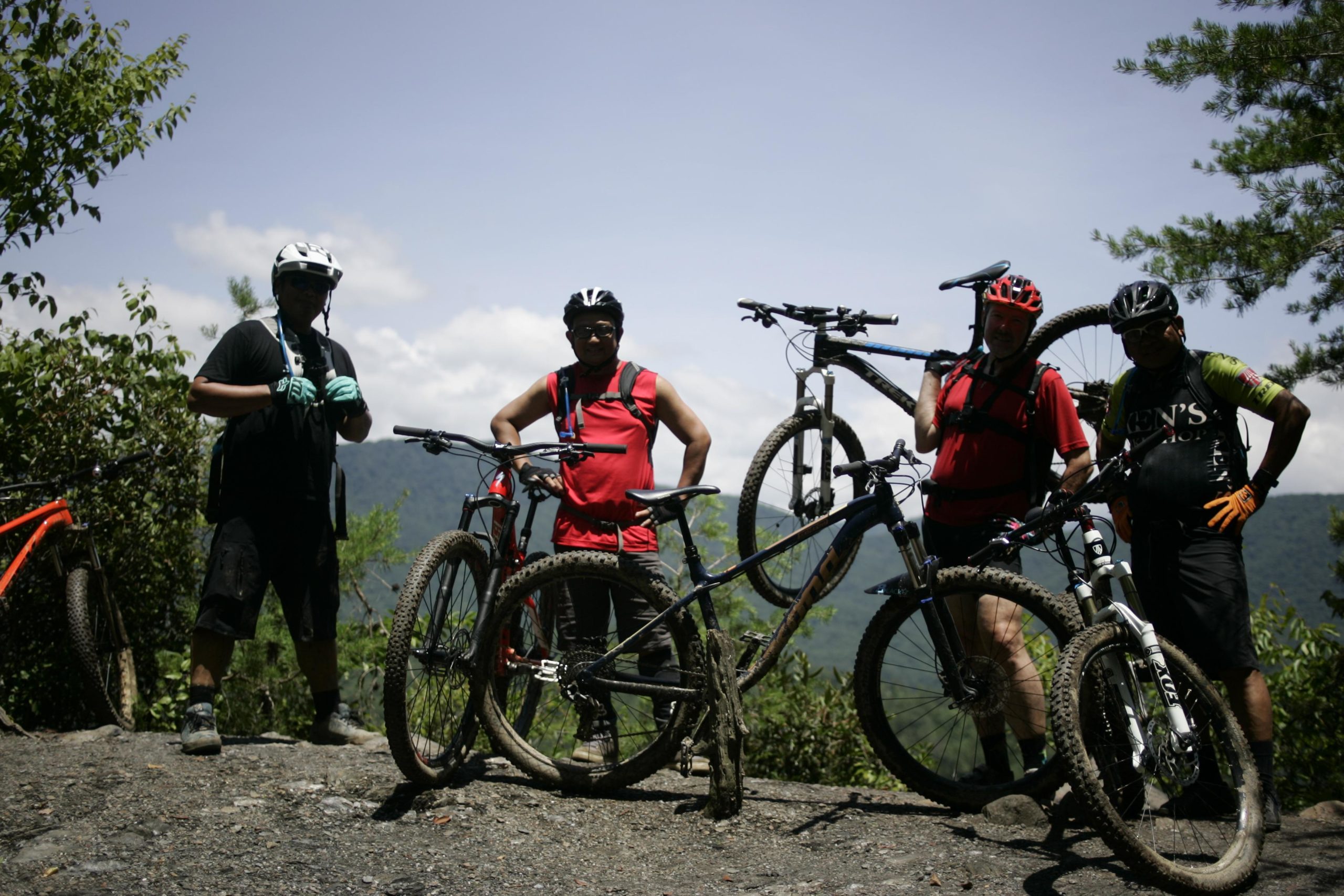 Four mountain bikers pose for a photo on a rocky trail, each holding or standing next to their bikes. They are wearing helmets and cycling gear, with a backdrop of green trees and distant mountains under a clear blue sky. Tsali Left Loop mountain bike trail.