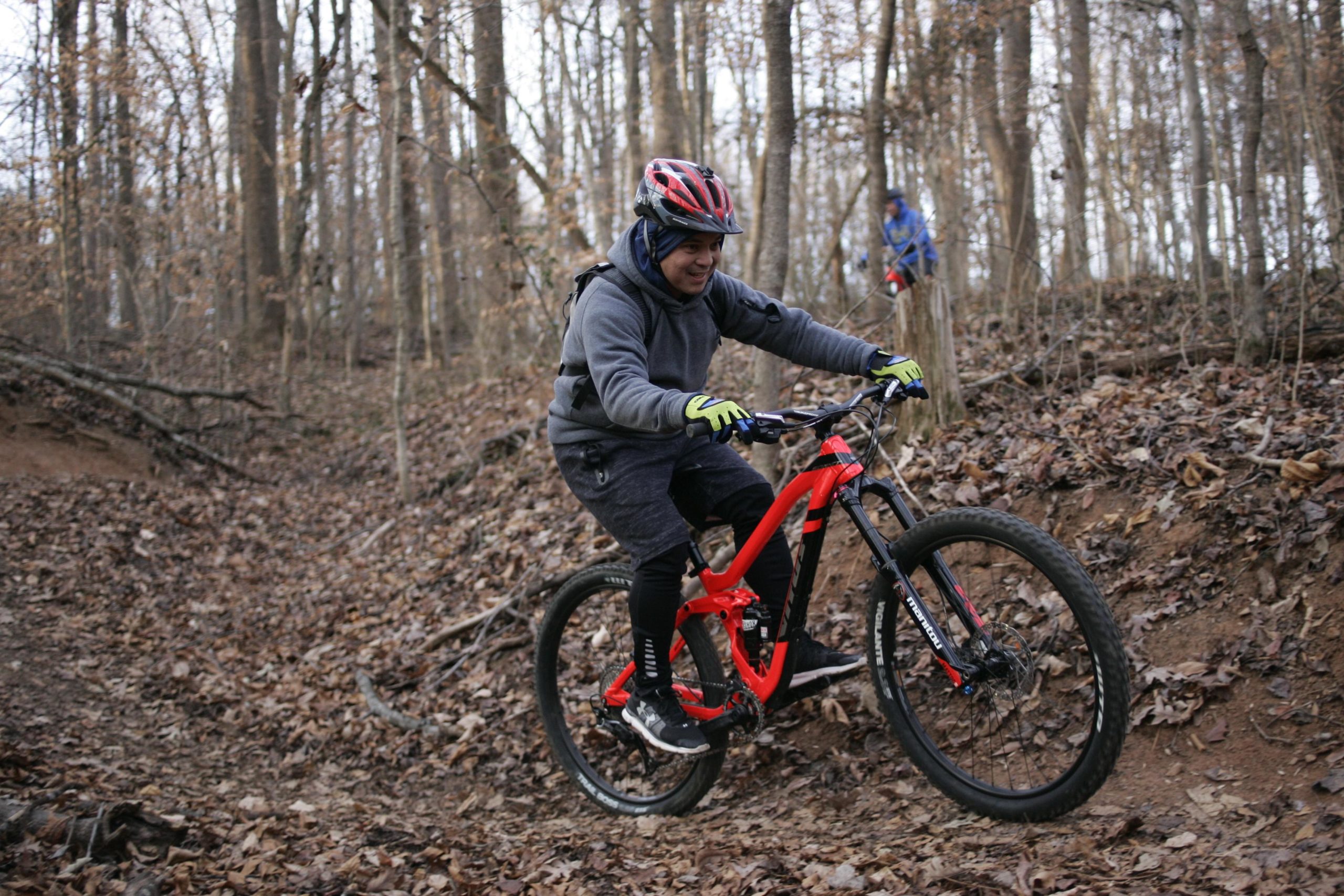 A mountain biker navigating a leaf-covered trail in a forest. The rider, wearing a helmet, gray hoodie, and gloves, is in an active pose, perched on a bright red mountain bike. Trees with bare branches surround the trail, indicating a late autumn or early winter scene. A blurred figure can be seen in the background. Hobby Park mountain bike trail.
