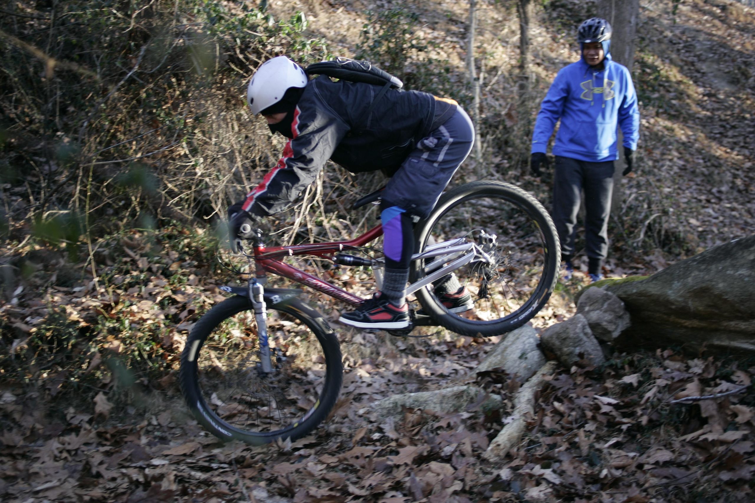 A person in winter cycling gear jumps a mountain bike over a rocky trail while another person watches from the side, surrounded by autumn leaves and trees. Hobby Park mountain bike trail.