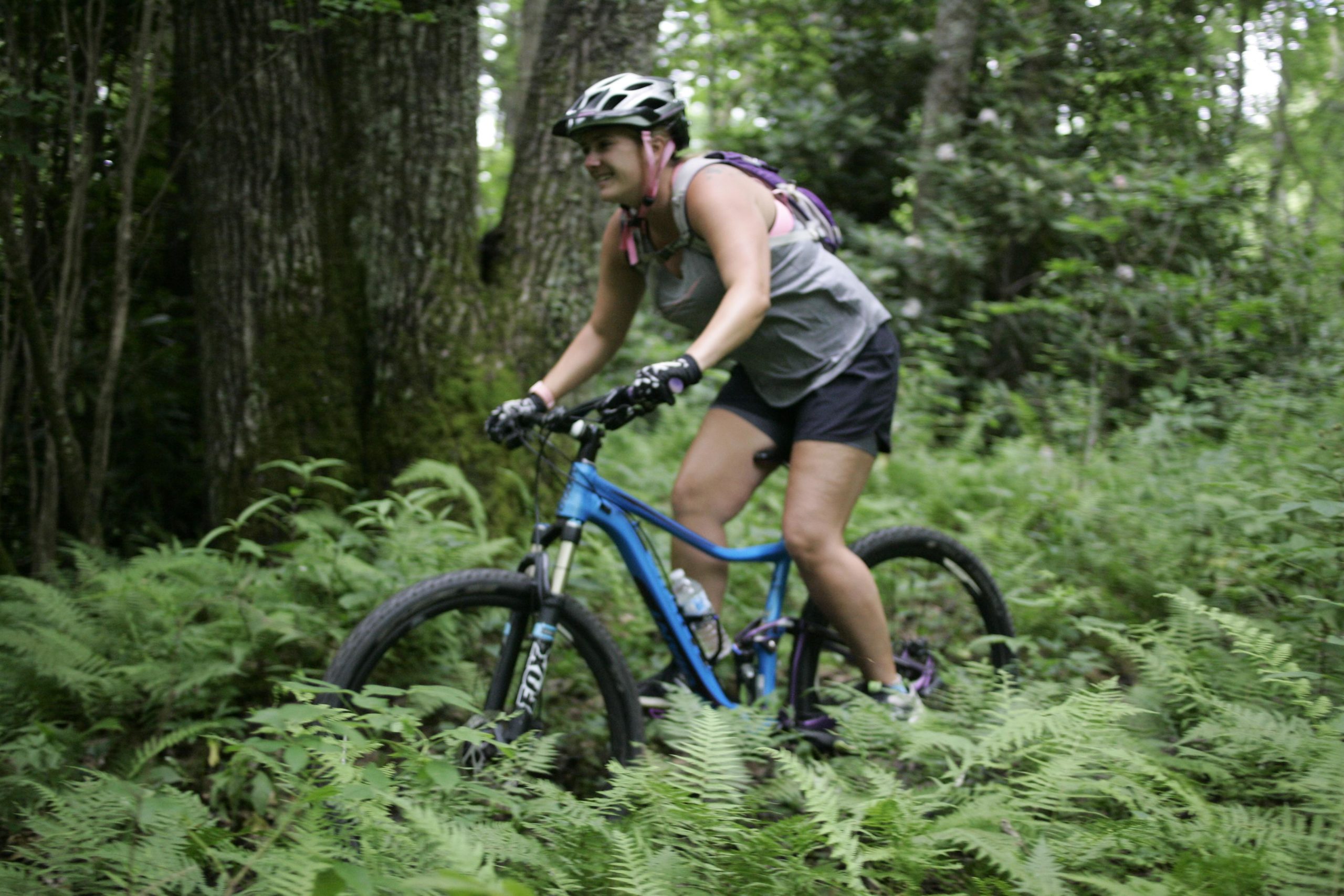 A woman riding a blue mountain bike through a lush, green forest trail surrounded by ferns and trees. She is wearing a gray tank top and shorts, along with a helmet for safety. Table Rock mountain bike trail.