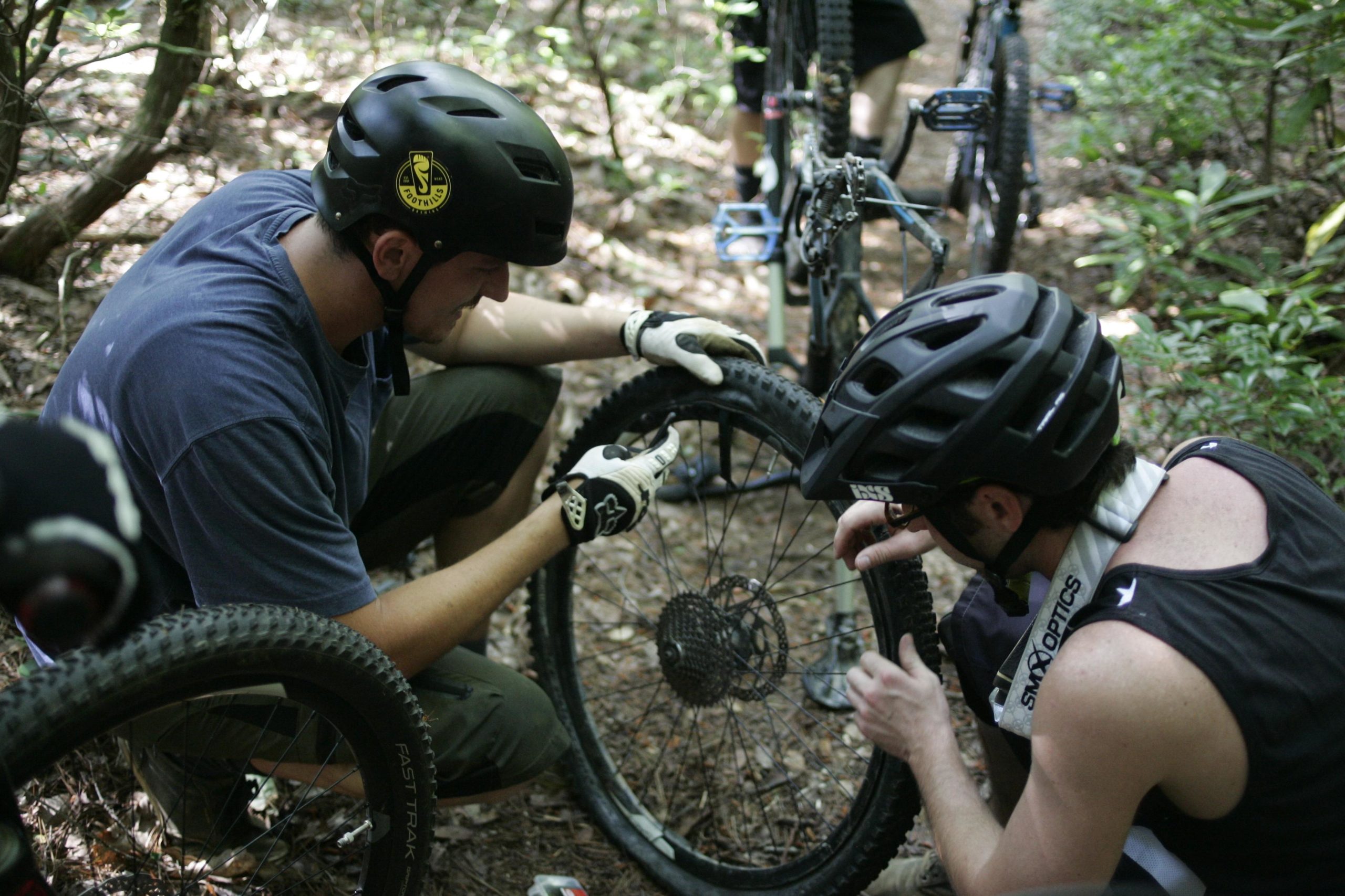 Two individuals in mountain biking gear are repairing a bike tire in a wooded area. One person is crouched down, examining the tire closely, while the other assists in the repair. The scene captures a moment of outdoor teamwork amid trees and natural surroundings. Table Rock mountain bike trail.