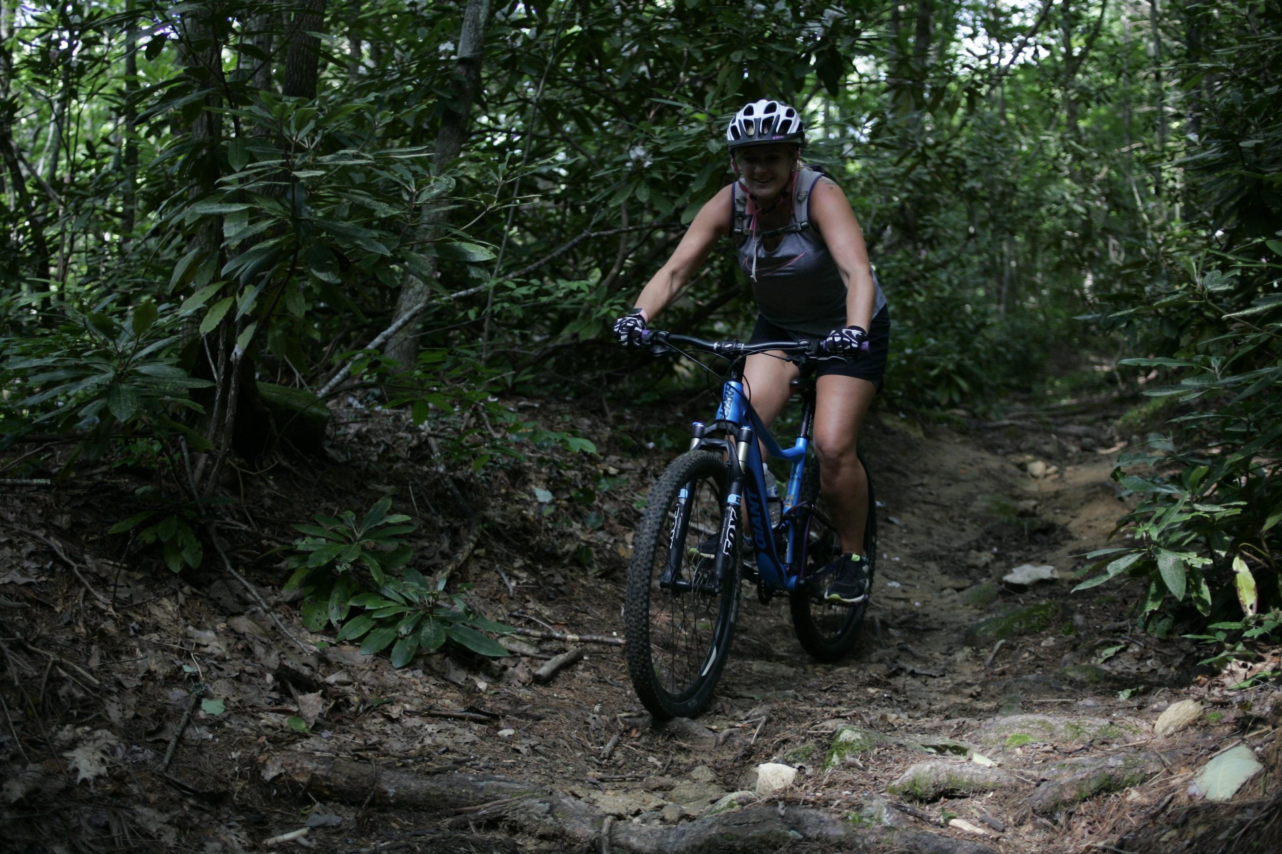 A woman riding a mountain bike on a narrow dirt trail surrounded by dense greenery in a forested area. She is wearing a helmet and gloves, appearing focused and engaged in biking on the rugged terrain. Table Rock mountain bike trail.