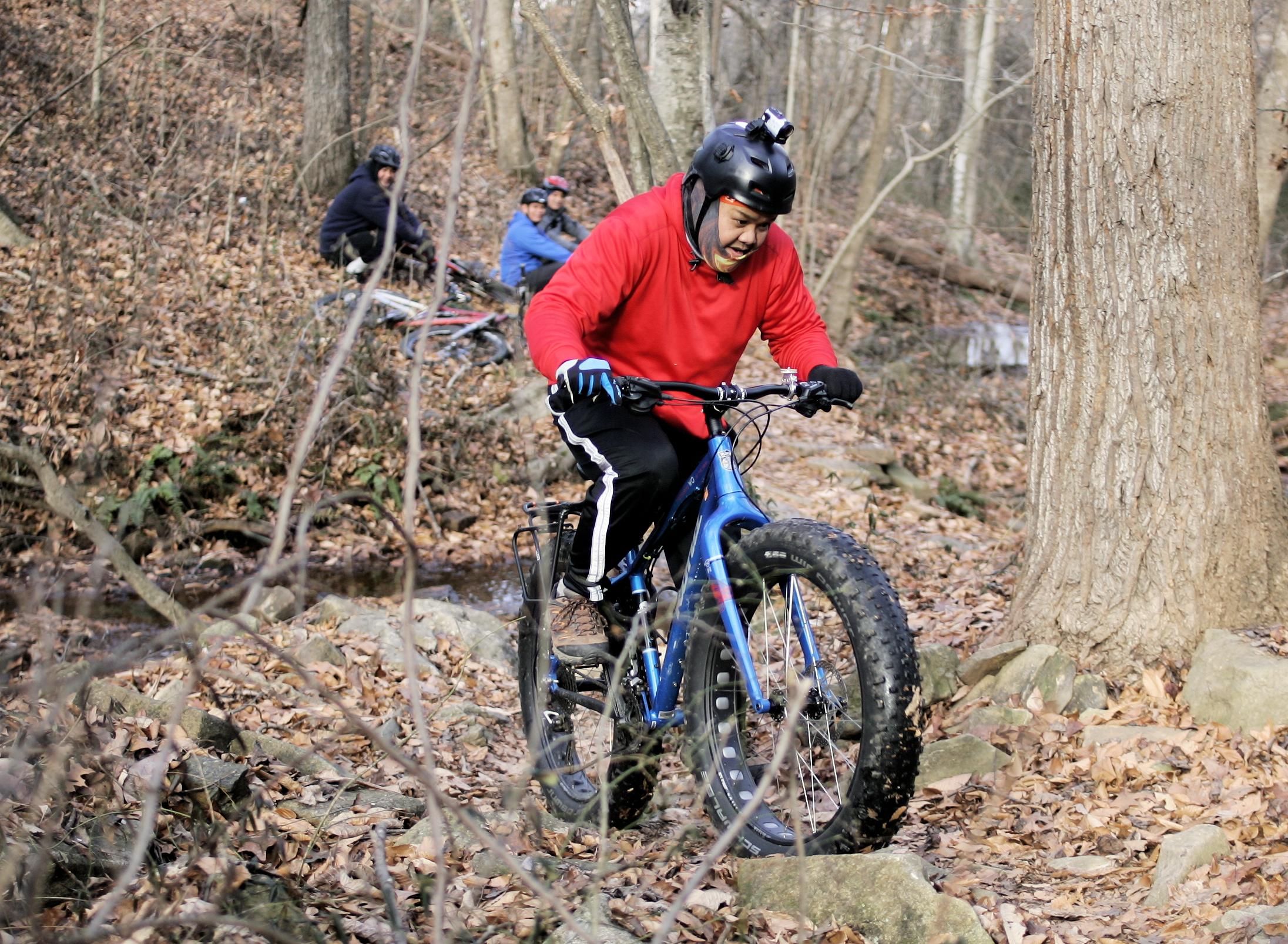 A person in a red sweatshirt and black pants rides a blue fat bike over rocky terrain covered with autumn leaves in a wooded area. Two other cyclists are visible in the background, among trees and fallen foliage. Hobby Park mountain bike trail.