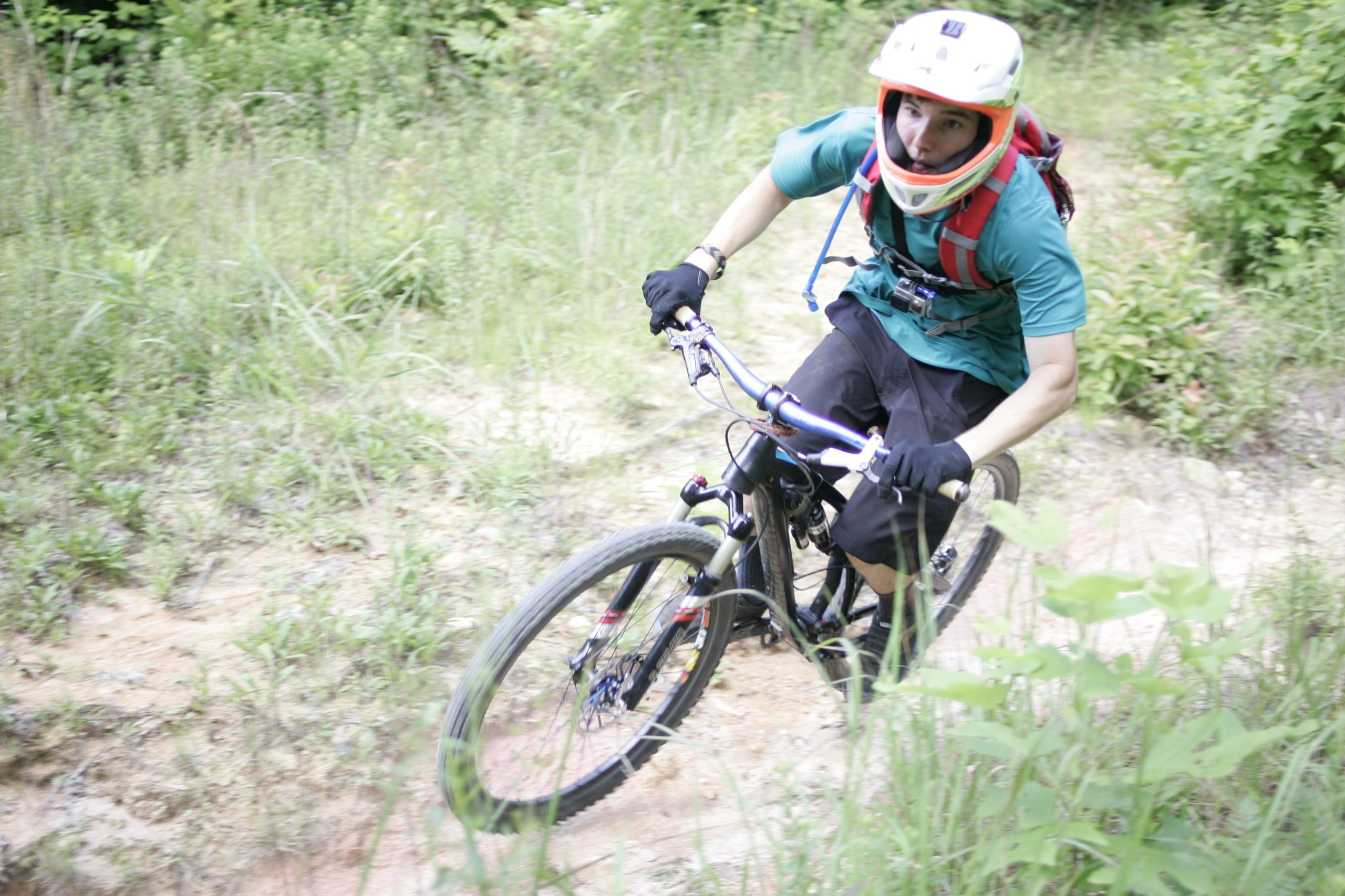 A mountain biker ascending a narrow, muddy trail surrounded by lush greenery. The rider is wearing a helmet and gloves, with a backpack and protective gear, showcasing an active outdoor adventure. Table Rock mountain bike trail.