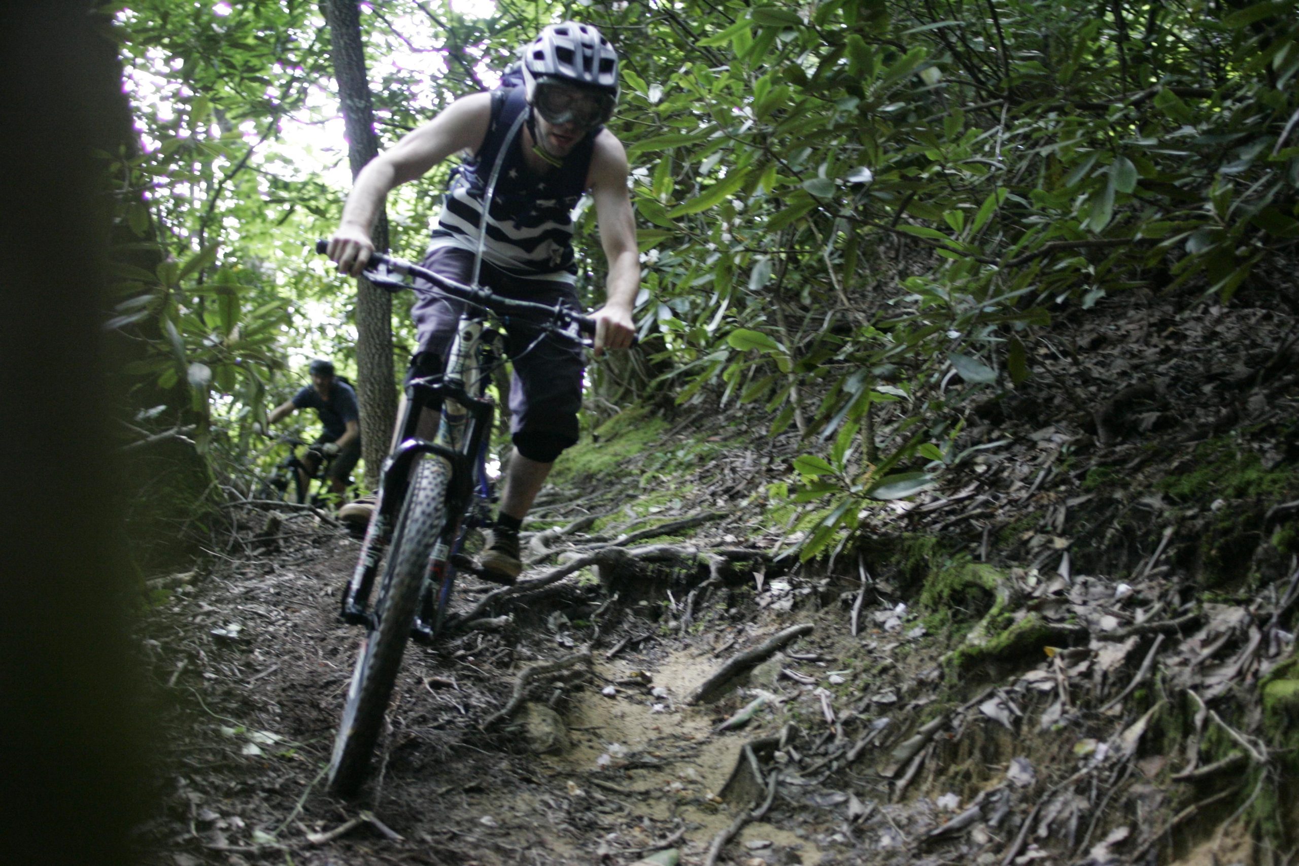A mountain biker navigating a rocky trail surrounded by dense foliage, with another cyclist in the background. The biker is wearing a helmet and protective gear, focused on maneuvering through the challenging terrain. Table Rock mountain bike trail.