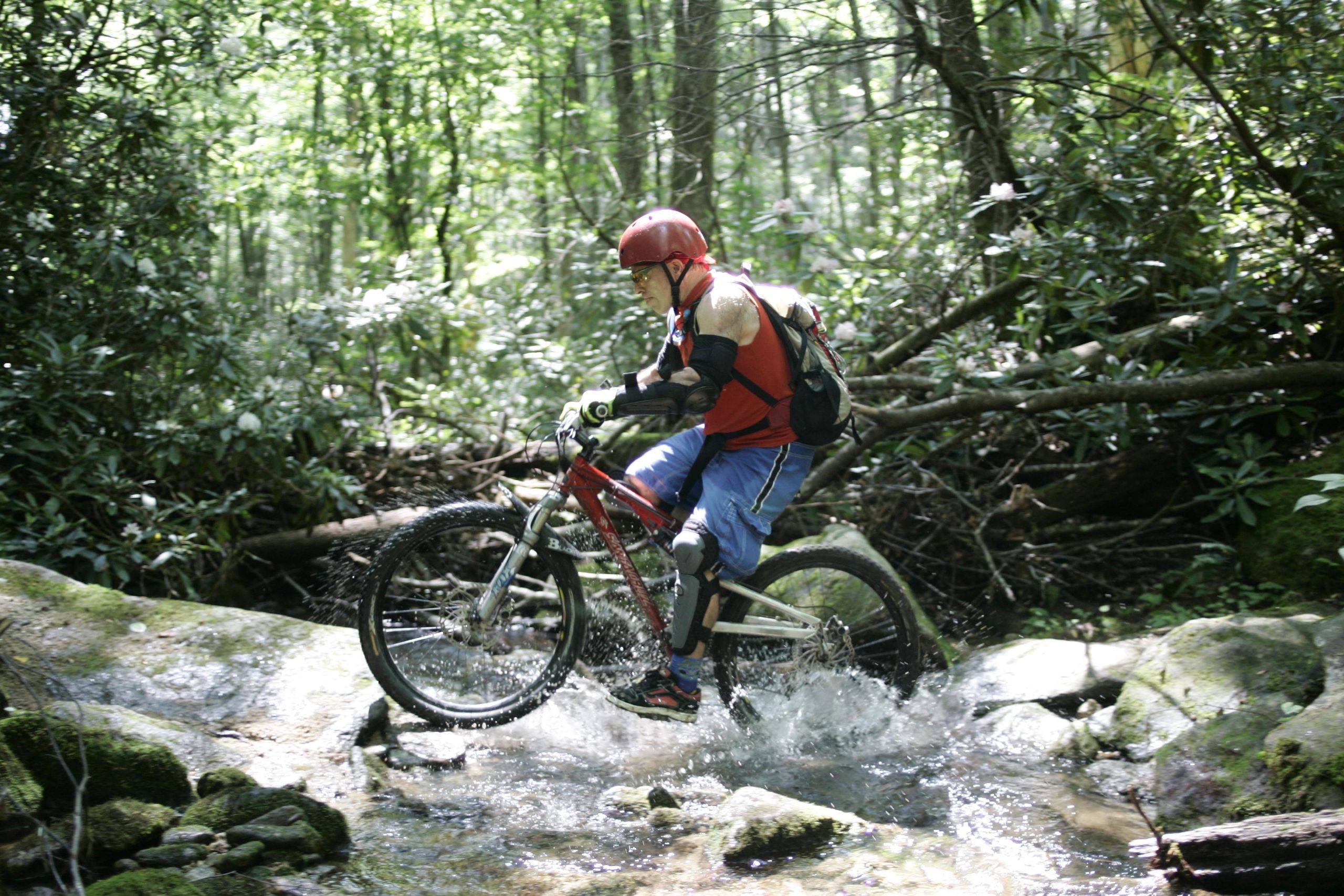 A mountain biker in a red helmet and red sleeveless shirt navigates through a rocky stream surrounded by dense greenery. Water splashes as the bike's wheel passes over the rocks, highlighting the adventure of off-road cycling in a natural setting. Table Rock mountain bike trail.