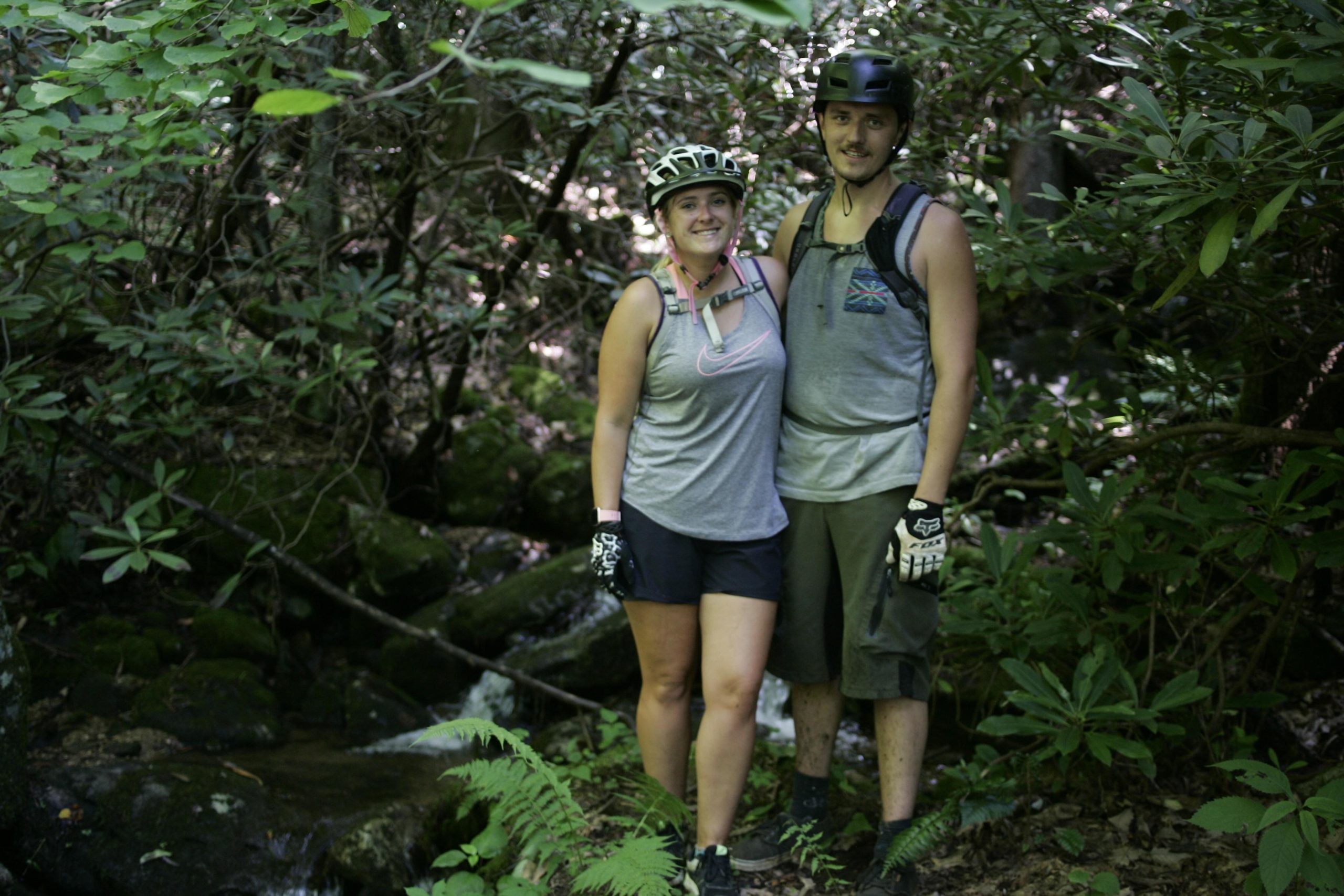 A couple stands together in a lush green forest, wearing mountain biking gear and helmets. They are smiling and posed in front of a small stream surrounded by rocks and vegetation. The sunlight filters through the trees, illuminating their relaxed demeanor in a natural outdoor setting. Table Rock mountain bike trail.