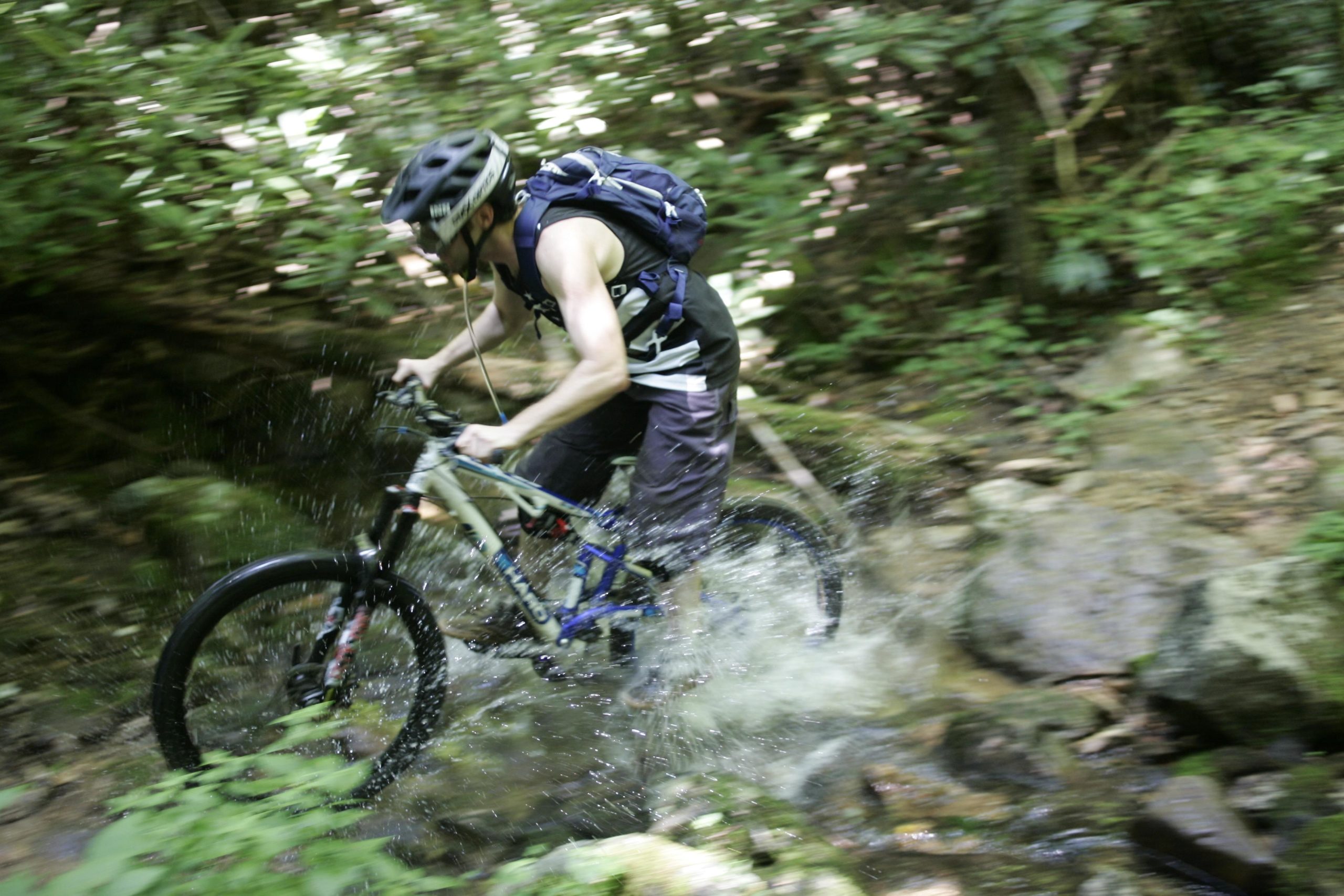 A mountain biker navigating through a stream, splashing water as he rides over rocky terrain surrounded by lush greenery. The cyclist is wearing a helmet and a backpack, demonstrating an active and adventurous outdoor activity. Table Rock mountain bike trail.