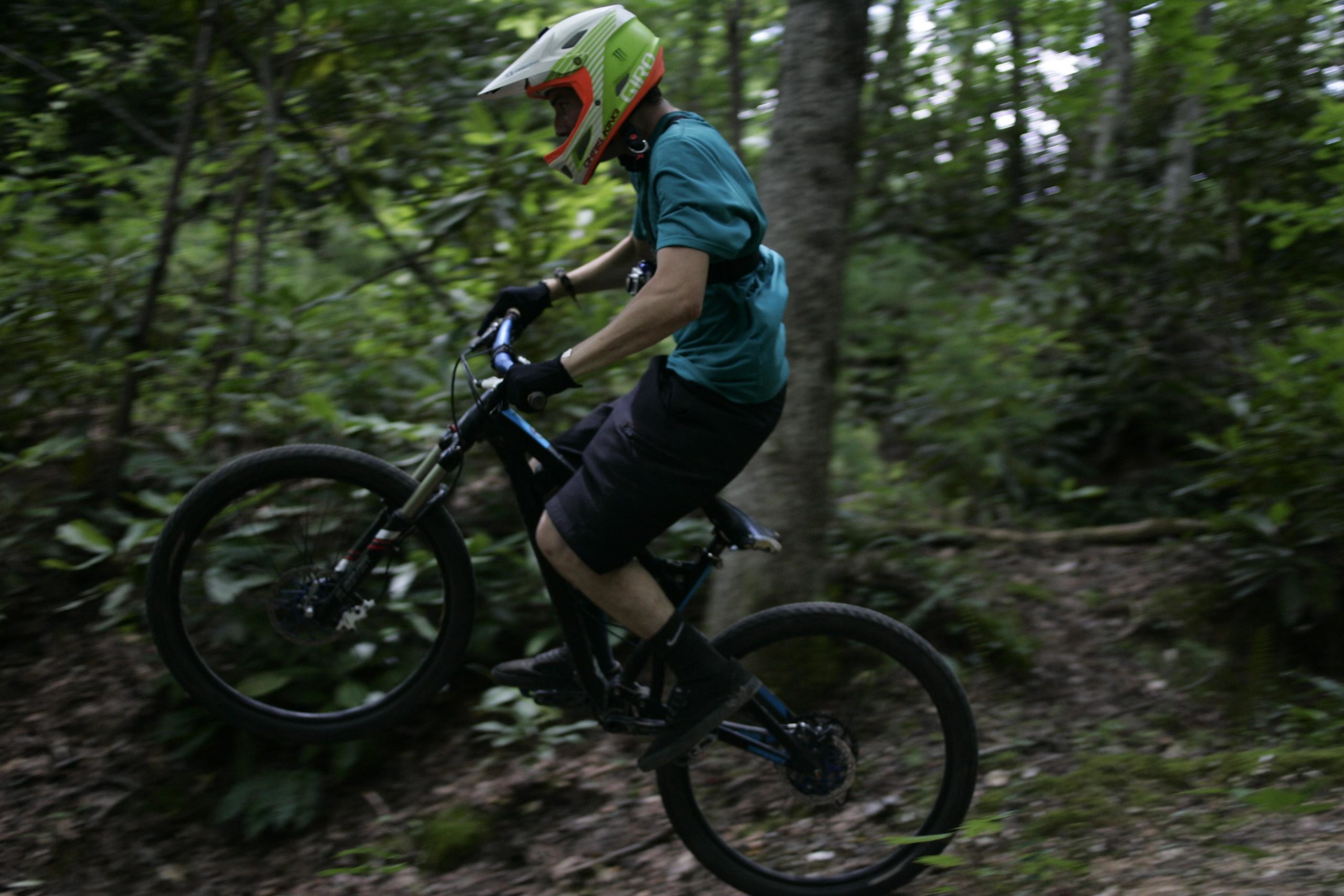 A mountain biker performing a jump on a dirt trail surrounded by greenery, wearing a helmet and protective gear. The biker is caught mid-air, showcasing an active and adventurous moment in a forested area. Table Rock mountain bike trail.