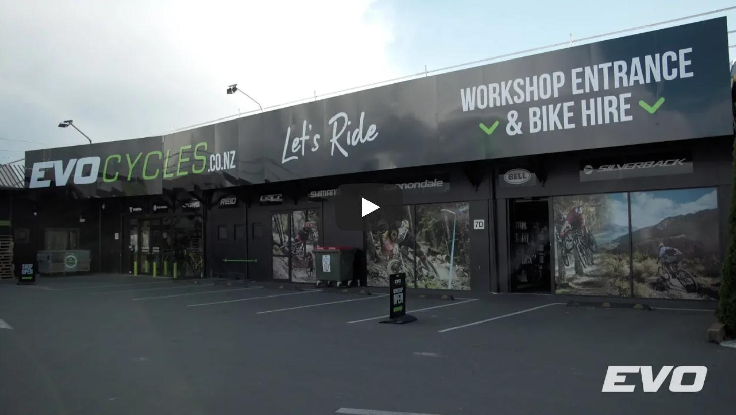 Image of the exterior of EVO Cycles, featuring a large sign with the company name and slogan "Let's Ride." The storefront has a workshop entrance and bike hire services indicated. The facade showcases images of cyclists and various bicycle brands. A sign states "Shop Open," and the parking area is visible in front of the store.
