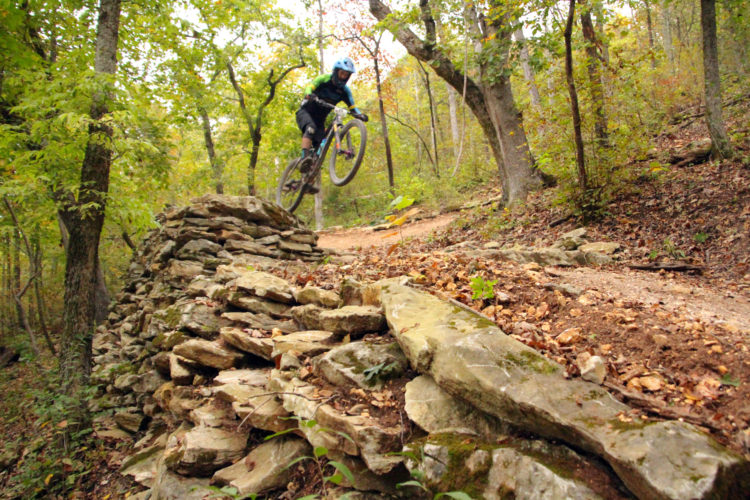 A mountain biker in motion leaps off a rocky ramp on a dirt trail surrounded by trees with autumn foliage. The bike is airborne, displaying a dynamic action shot that captures the excitement of outdoor biking.