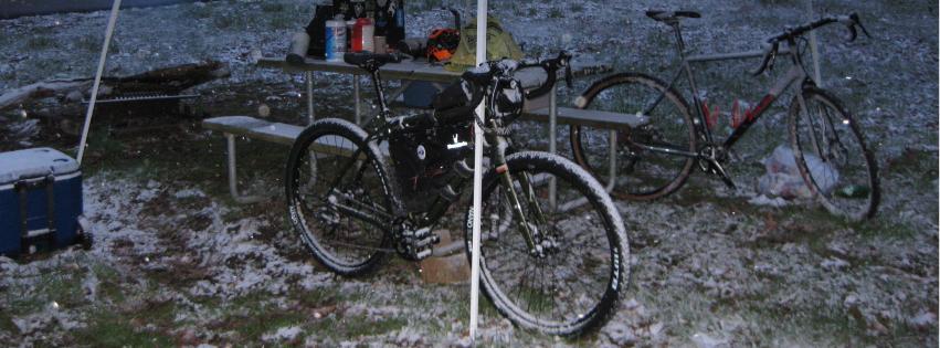 Salsa Jouneryman: A camping scene featuring two bicycles parked near a picnic table under a tent. The ground is covered with a light dusting of snow, and there are supplies visible on the table, including containers and gear. The setting appears to be outdoors in a winter environment.