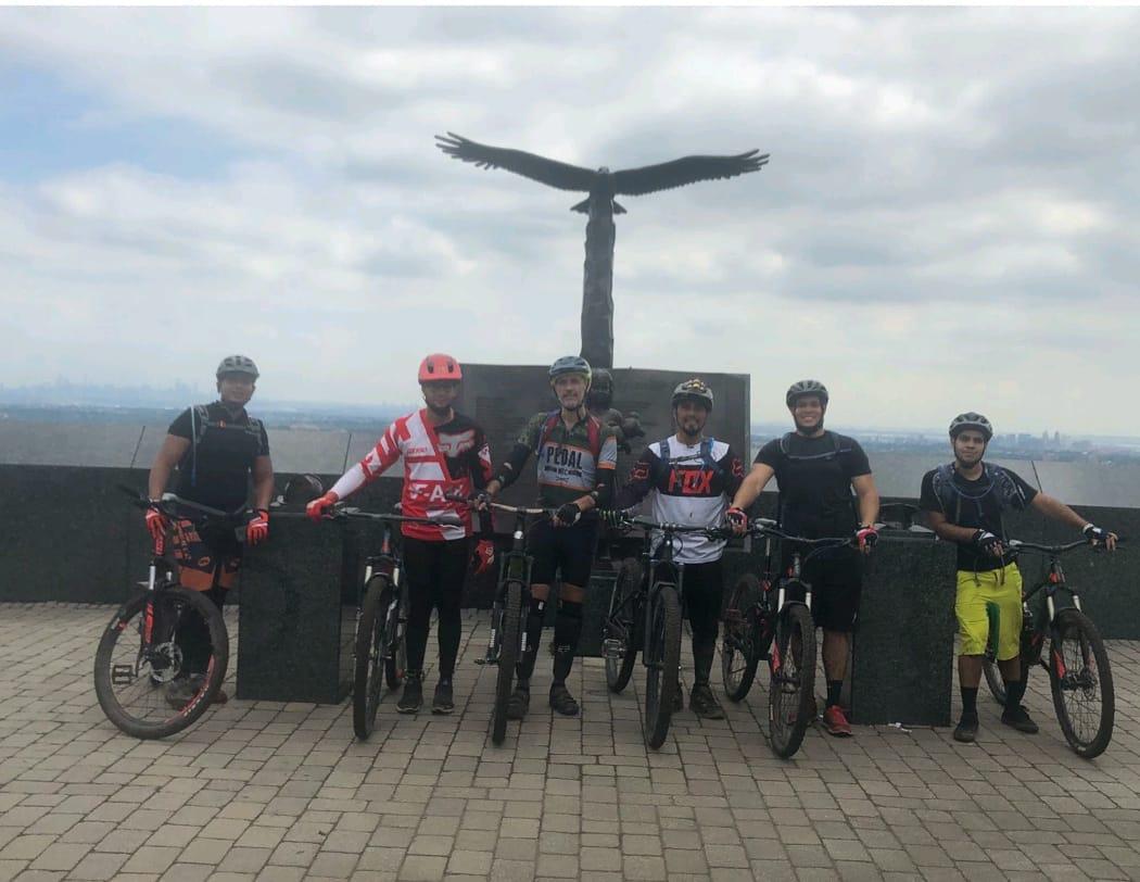 A group of six mountain bikers pose for a photo at a lookout point featuring a large eagle statue. They are wearing cycling helmets and gear, with their bicycles beside them. The backdrop includes a cloudy sky and a view of a distant city skyline. The scene conveys a sense of camaraderie and adventure in a scenic outdoor location. Hilltop reservation mountain bike trail.