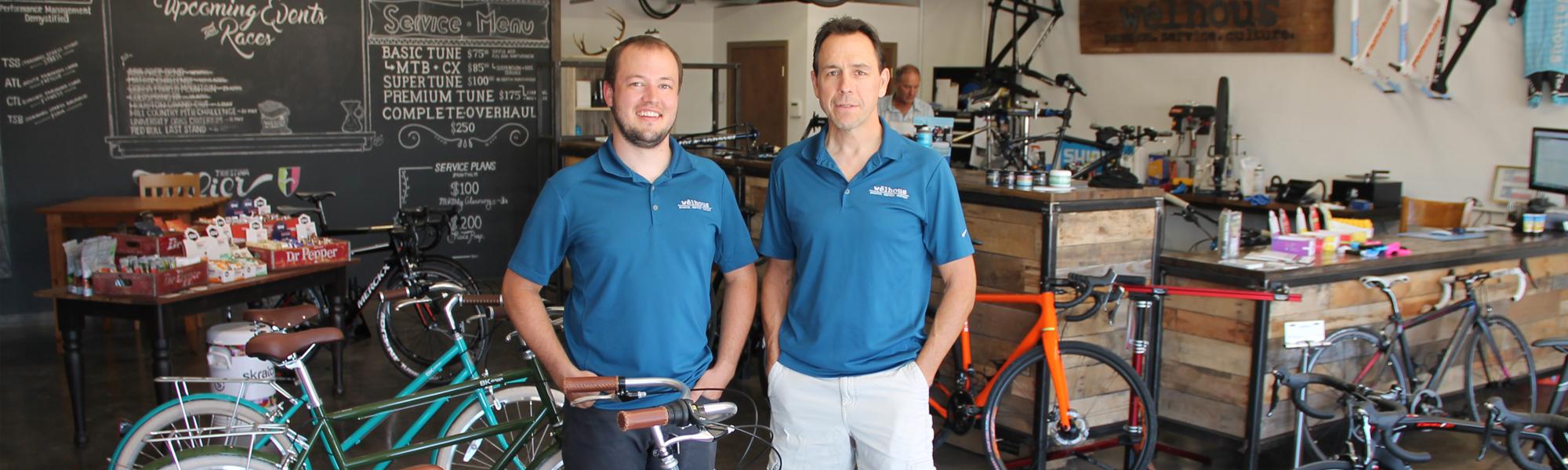 Two men stand in a bicycle shop, both wearing blue polo shirts with a logo. They are positioned in front of a wooden counter filled with bike maintenance supplies. In the background, various bicycles are displayed, alongside a chalkboard listing upcoming events and service options. The interior has a modern, rustic feel with wooden accents.