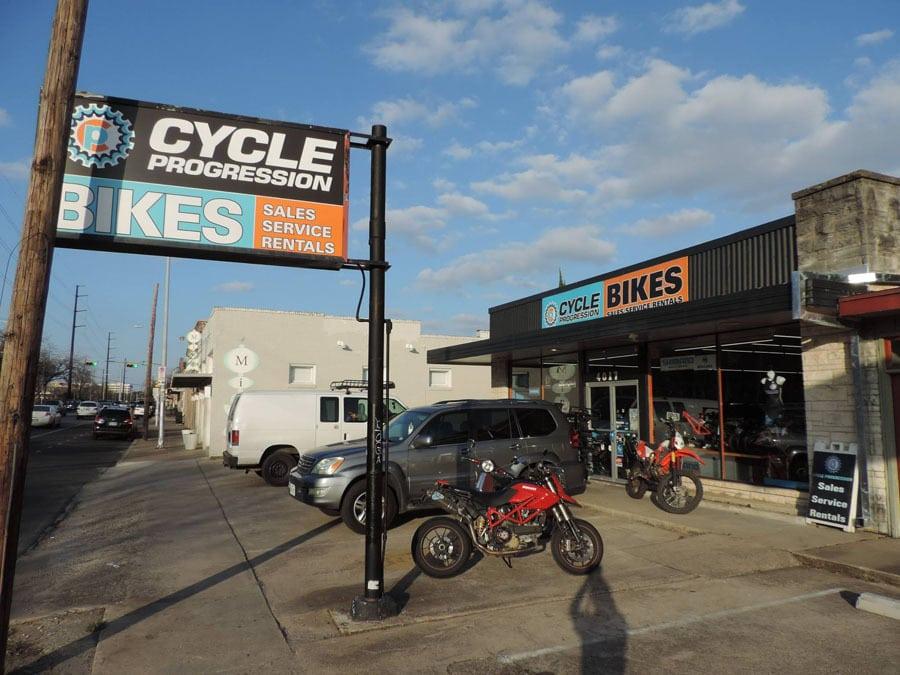 A storefront for "Cycle Progression" featuring a large sign that reads "CYCLE" and "BIKES" indicating sales, service, and rentals. The building displays various motorcycles both outside and visible through the windows, with parked vehicles alongside. The scene is set against a bright blue sky with scattered clouds.