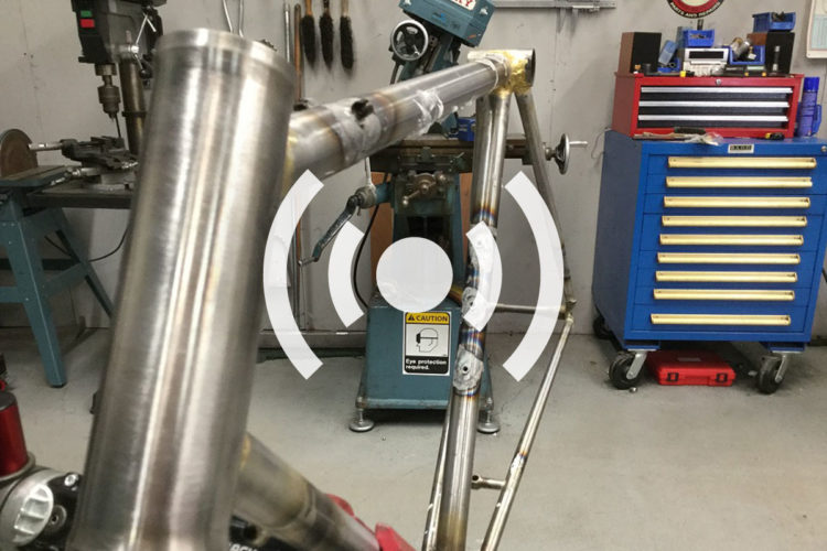 A close-up view of a metal bicycle frame being crafted, showing polished and welded joints. In the background, a drill press and a toolbox with multiple drawers are visible, along with various tools hanging on the wall. A caution sign about eye protection is also seen. The image includes a graphic element resembling sound waves over the frame.