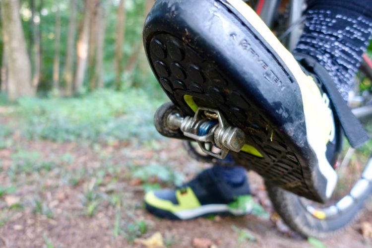 Close-up view of a mountain bike shoe with a cleat, resting on a forest floor. The shoe features a black sole with textured grip and bright yellow accents. In the background, greenery and trees are visible, indicating an outdoor cycling environment.