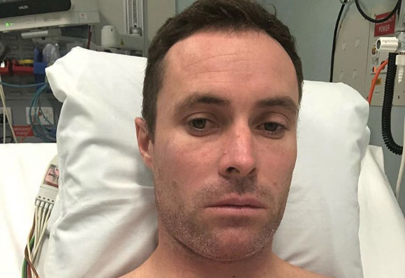 A man with short dark hair is lying on a hospital bed, looking directly at the camera with a serious expression. He has a stubble beard and is wearing a hospital gown. Medical equipment and supplies are visible in the background.