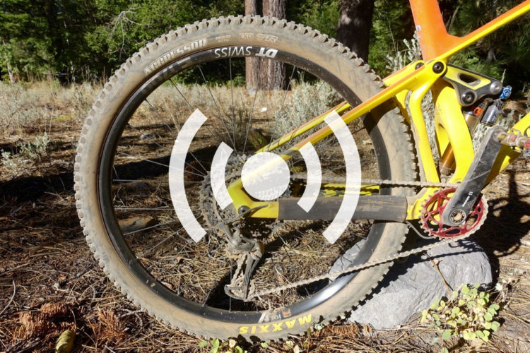 A close-up view of a mountain bike's rear wheel and drivetrain, featuring a yellow frame, a black Maxxis tire, and visible components like the derailleur and chain. The background shows a natural setting with plants and trees. A graphic overlay of sound waves is present in the center of the image.
