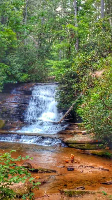 A serene waterfall cascading over rocky steps into a shallow stream, surrounded by lush green foliage and trees in a tranquil forest setting. Stonewall Falls Loop mountain bike trail.
