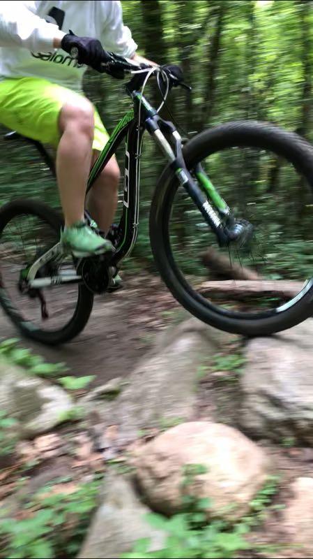 A mountain biker in mid-jump over rocky terrain on a forest trail. The cyclist is wearing a white long-sleeve shirt and bright yellow shorts, with green shoes visible. The bike features black and green accents as it navigates a rugged path surrounded by lush greenery. Lebanon Hills mountain bike trail.