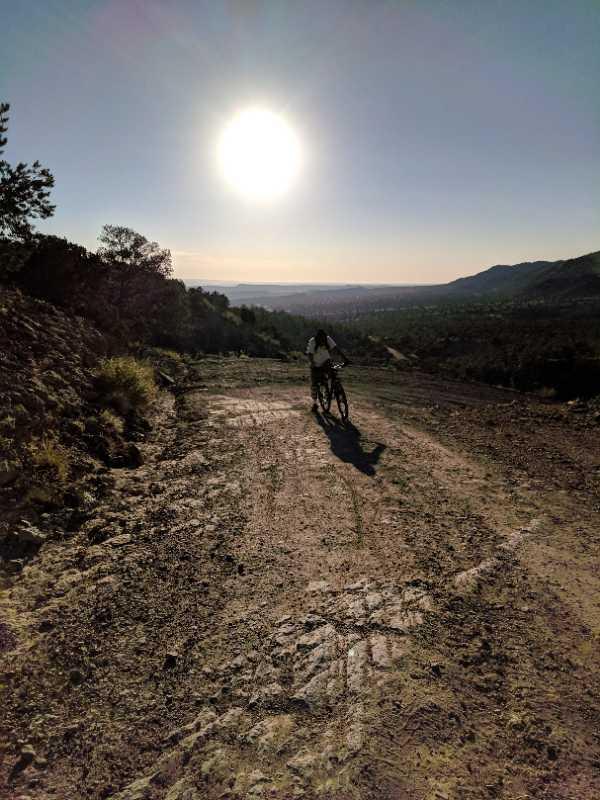 A silhouette of a person riding a bicycle on a dirt path, with the sun hanging low in the sky, casting light over a valley and distant mountains. Trees are visible on the left side of the image, and the landscape fades into a hazy horizon. The scene conveys a sense of adventure and outdoor exploration. South Canon Trails mountain bike trail.
