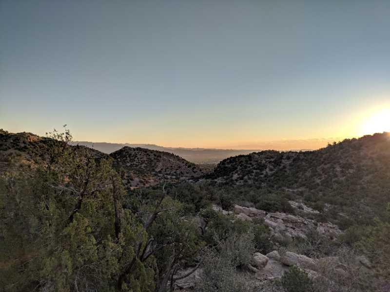 A panoramic view of rolling hills and mountains during sunset, with a clear sky transitioning to twilight. Sparse vegetation, including some trees and rocky terrain, is visible in the foreground, while the sun casts a warm glow on the landscape. South Canon Trails mountain bike trail.