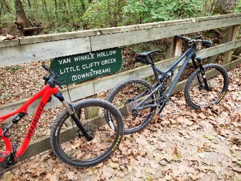 Two mountain bikes, one red and one black, are parked beside a wooden sign indicating "Van Winkle Hollow Little Clifty Creek (Downstream)." The background features a wooded area with fallen leaves on the ground. Hobbs State Park Conservation Area mountain bike trail.