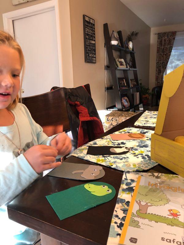 A young child is seated at a table, engaging in a craft activity. The child is working with colorful felt pieces, including a green frog and other animal shapes, while focused on a project. The table is decorated with a floral tablecloth, and in the background, there are shelves with books and decorations. A craft box and an illustrated guidebook are also visible. Cape Split mountain bike trail.