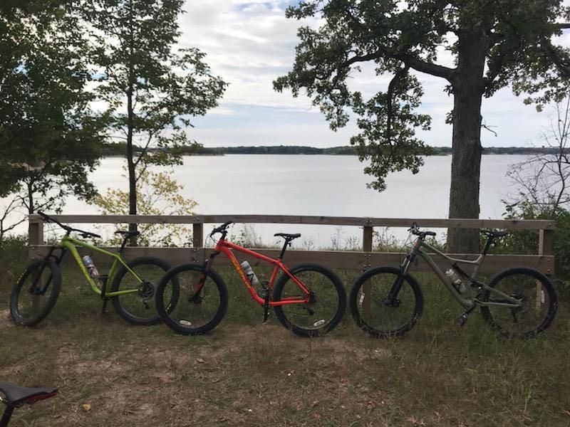 Three mountain bikes in green, orange, and gray parked near a wooden railing overlooking a calm lake, surrounded by trees and grassy land. The sky is partly cloudy, suggesting a serene outdoor setting. Camp Camfield mountain bike trail.