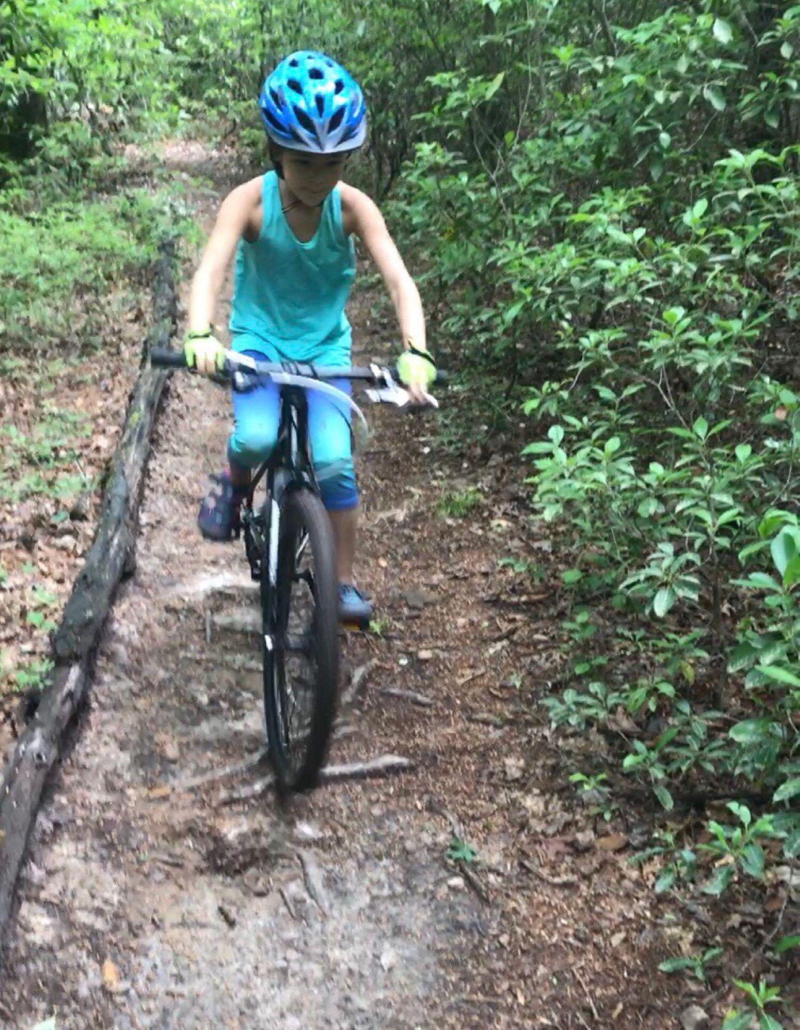 A child wearing a blue helmet and a turquoise outfit rides a bicycle along a dirt path surrounded by greenery. The child appears focused and balanced as they navigate the uneven terrain. Lower Sidehill / 137a mountain bike trail.