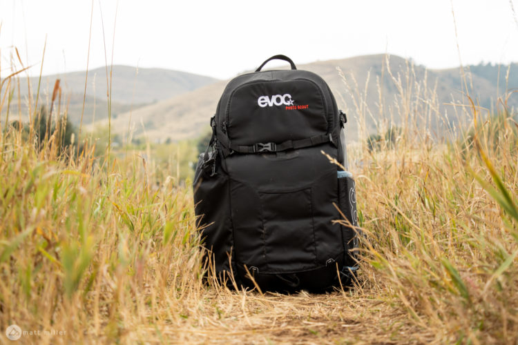 A black backpack labeled "evoc" is positioned in a field with tall grass, surrounded by rolling hills in the background under a cloudy sky.