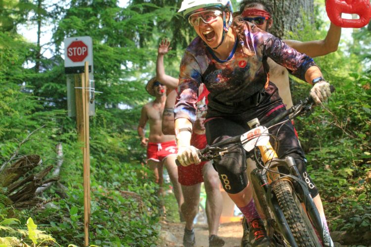 A mountain biker wearing a colorful jersey and helmet rides excitedly down a forest trail, looking back with a joyful expression. In the background, two enthusiastic spectators cheer her on, one wearing a hat and shorts, and the other holding a red object, with a "STOP" sign visible nearby. Lush greenery surrounds the scene, creating a vibrant outdoor atmosphere.