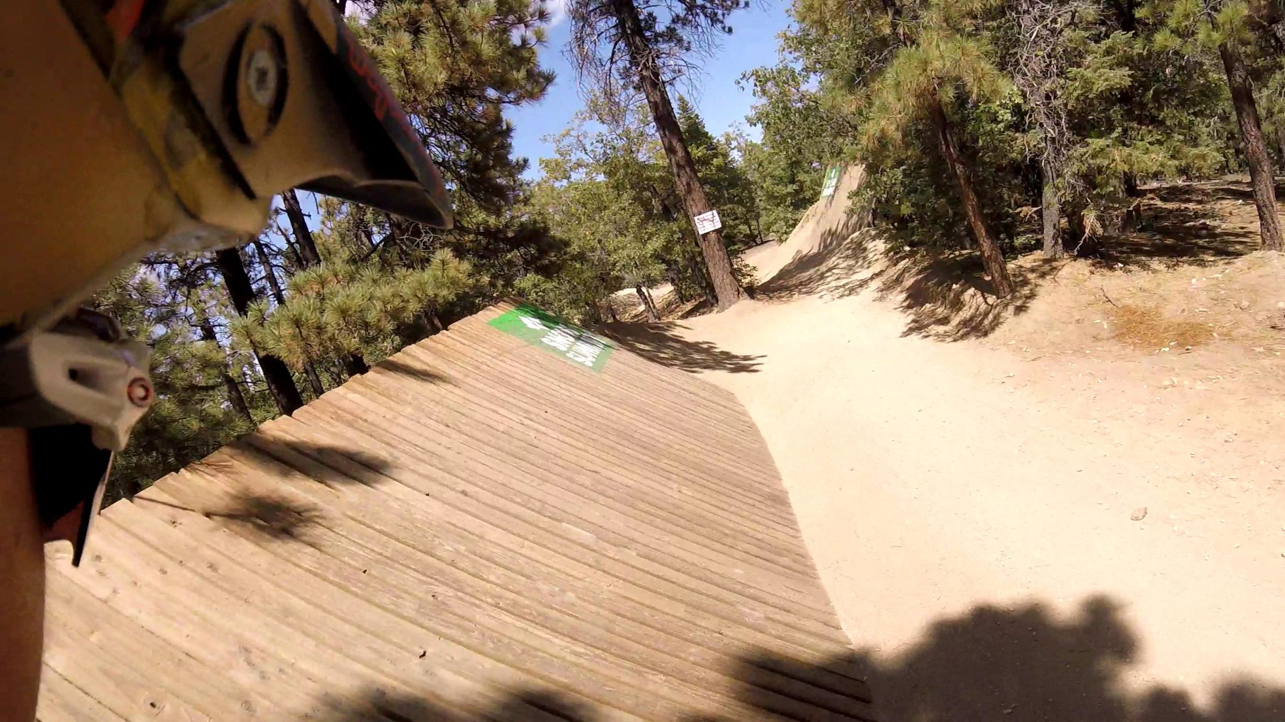 A close-up view of a dirt bike ramp in a forested area, with trees in the background and a clear blue sky. The angled wooden ramp is partially visible, showing a green sign. The image captures the perspective from a rider's helmet, suggesting an exciting biking experience. Big Bear Mountain Resort mountain bike trail.
