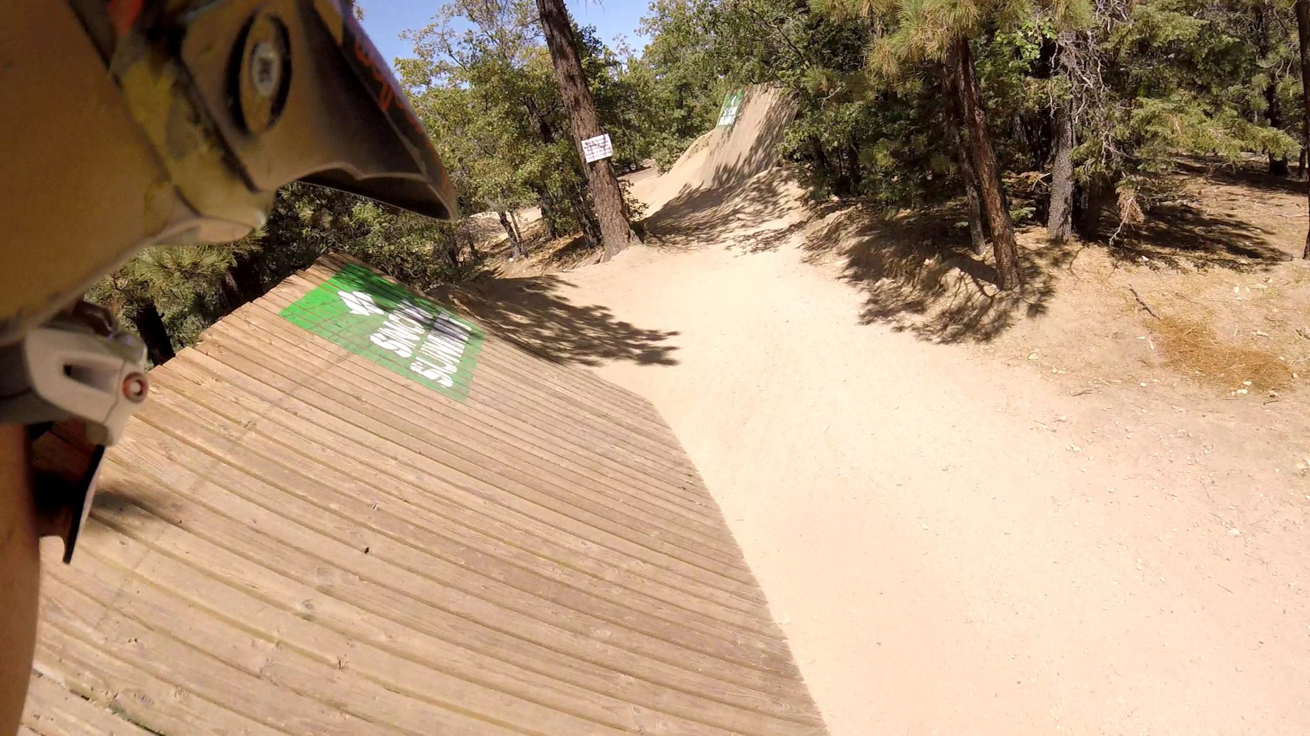 A close-up view from a mountain biker's perspective, featuring a wooden ramp and a sandy path winding through a forested area. The ramp has a green sign visible that reads "Caution!" and tall trees surround the scene, indicating an outdoor biking or trail area. Big Bear Mountain Resort mountain bike trail.