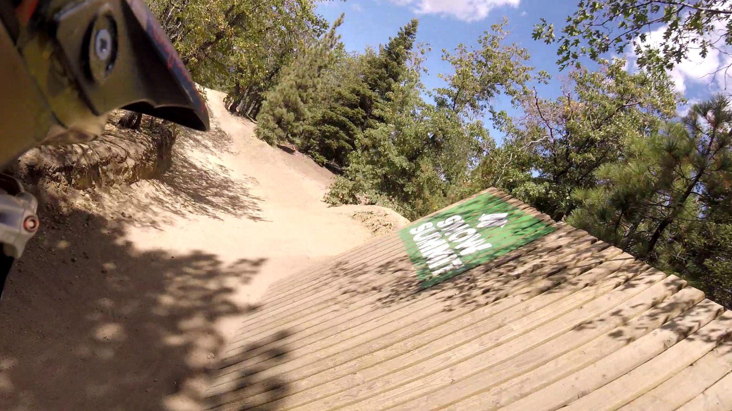 A close-up view of a sloped wooden ramp with a green sign that reads "Snow Summit" in white letters, surrounded by trees and a dirt path. The photo captures a sunny day, with blue sky and scattered clouds. The focus is on the ramp, suggesting an outdoor recreational area for biking or other activities. Big Bear Mountain Resort mountain bike trail.