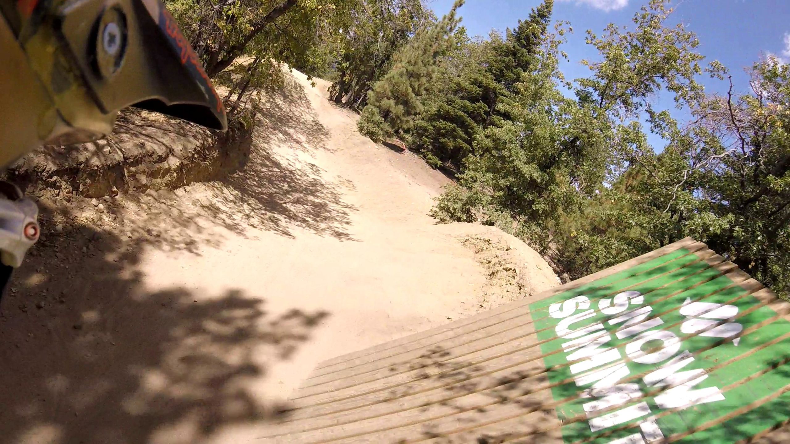 Image of a dirt path winding through a wooded area, with a partially visible green sign that reads "Snow Summit." The scene captures a sunny day with blue skies and light shadows from the surrounding trees, indicating a trail suited for outdoor activities like biking or hiking. Big Bear Mountain Resort mountain bike trail.