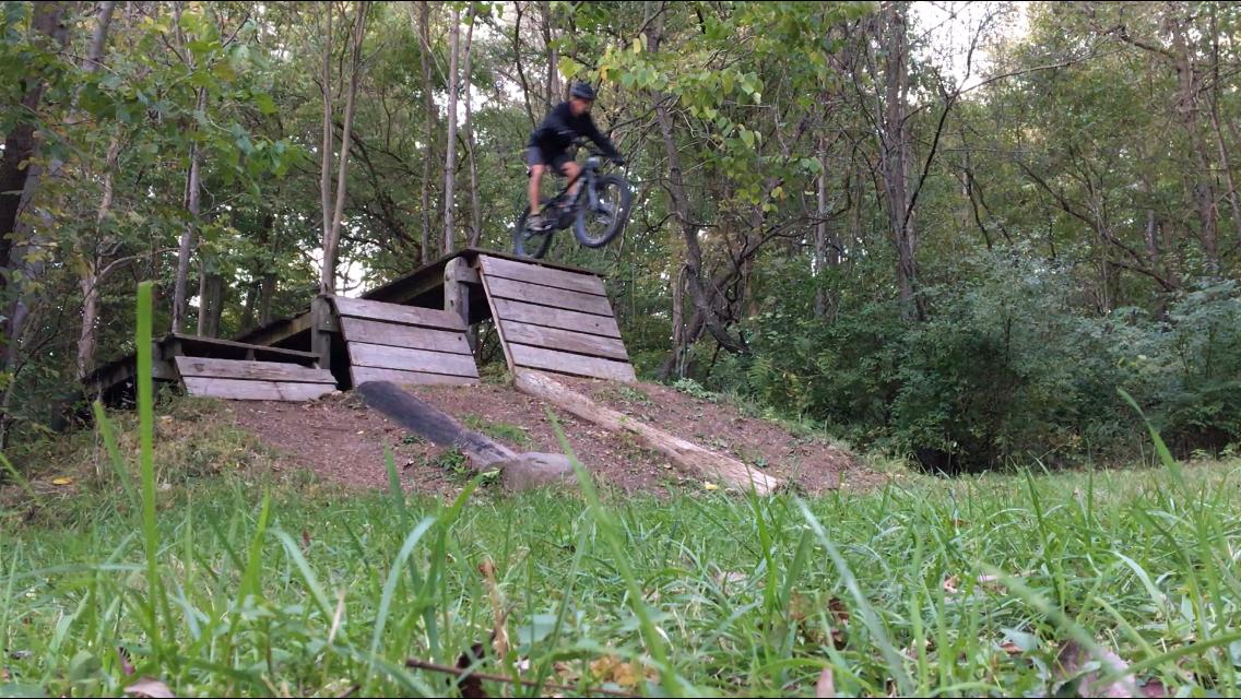 A cyclist in mid-air performing a jump off a wooden ramp in a wooded area, surrounded by grass and trees. Farmdale Reservoir Recreation Area mountain bike trail.