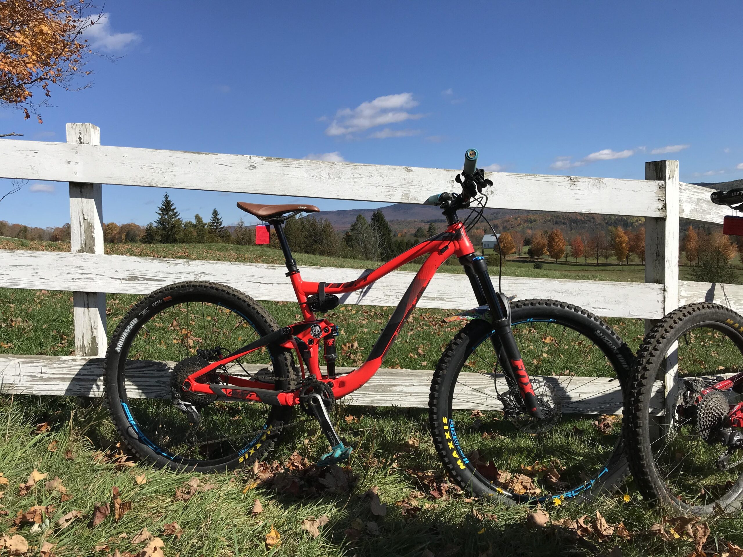 Giant Trance 27.5 2: A bright red mountain bike is leaning against a white wooden fence, set against a backdrop of blue sky and green fields. The ground is covered with fallen leaves, and trees with autumn foliage are visible in the distance.