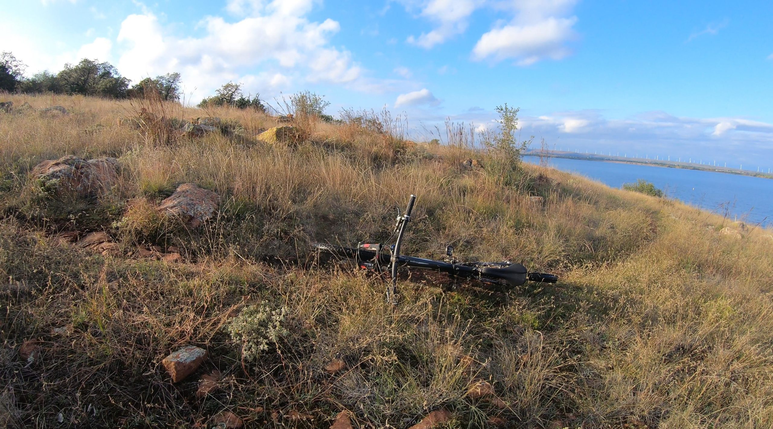 A black bike lying on the ground amidst tall grass and small rocks, with a scenic view of a lake and wind turbines in the distance under a partly cloudy sky. Lake Lawtonka Trails mountain bike trail.