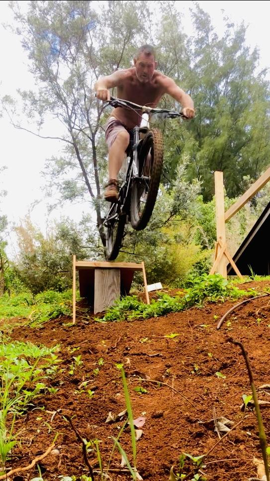YT Industries YT Jeffsy CF: A shirtless man riding a mountain bike performs a jump off a dirt ramp, surrounded by greenery and trees in a natural setting.