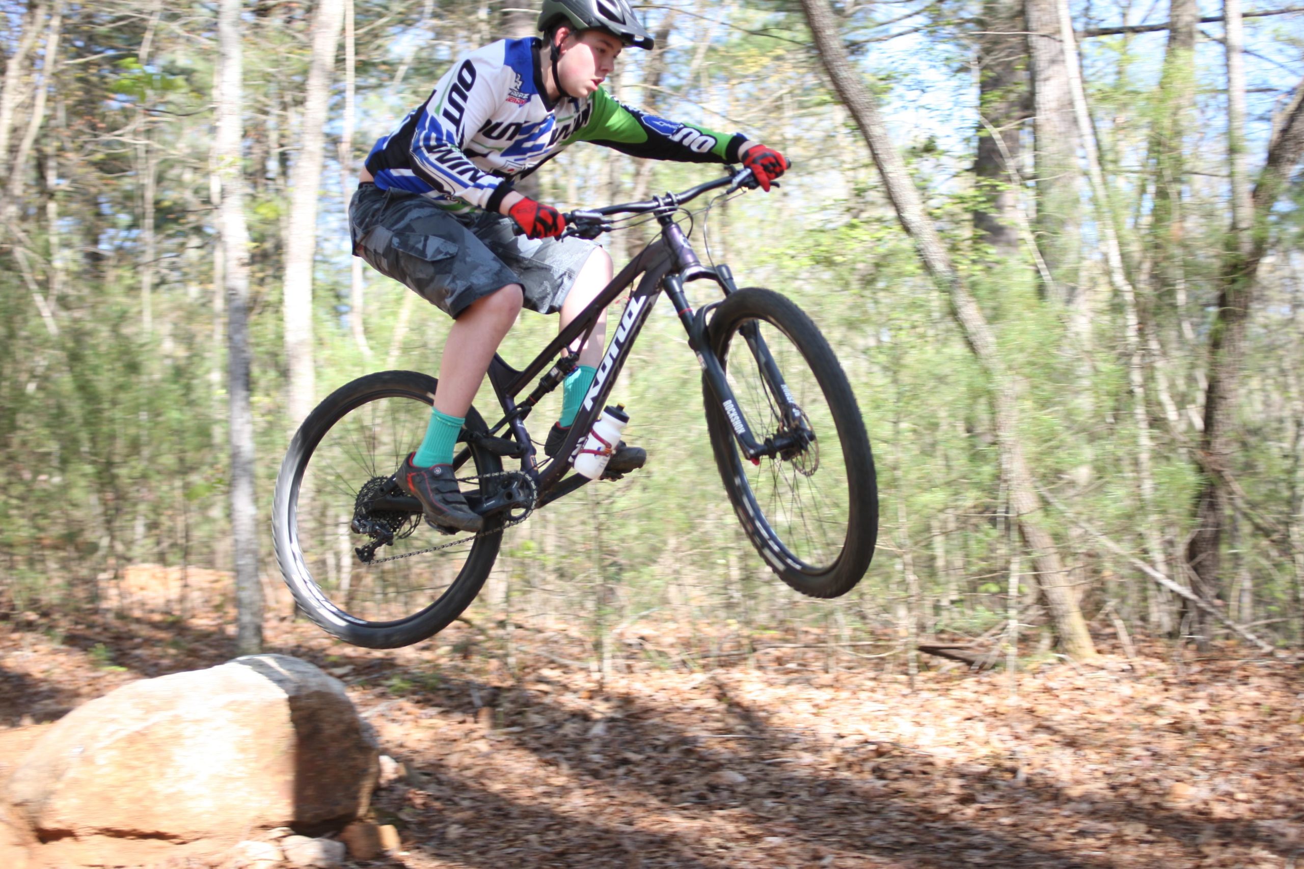 A young male cyclist wearing a helmet and protective gear jumps off a rock while riding a mountain bike on a wooded trail. Low shrubbery and fallen leaves surround the path, suggesting an outdoor adventure in natural surroundings. Dark Mountain Trail mountain bike trail.