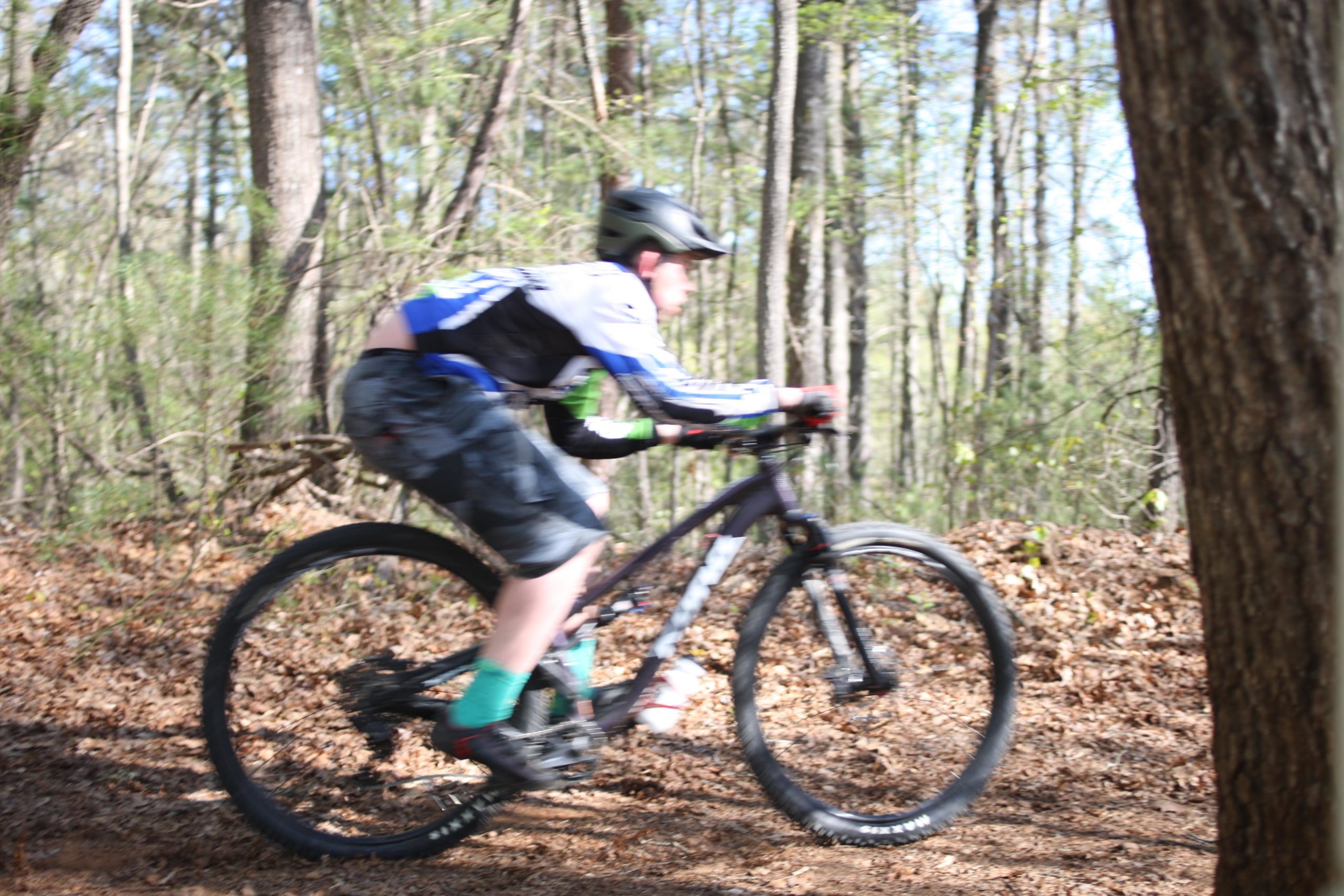 A person riding a mountain bike on a dirt trail in a wooded area, captured with a motion blur effect to emphasize speed. The cyclist is wearing a helmet and a jersey with blue and green accents, alongside shorts. Surrounding trees and fallen leaves create a natural outdoor setting. Dark Mountain Trail mountain bike trail.
